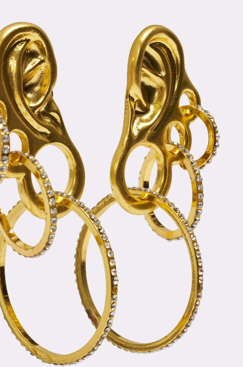 Close-up of the IN MY EAR EARRING: gold, ear-shaped earrings with a layered ear design, multiple rhinestone hoops, and small crystal accents, set against a plain white background. 3