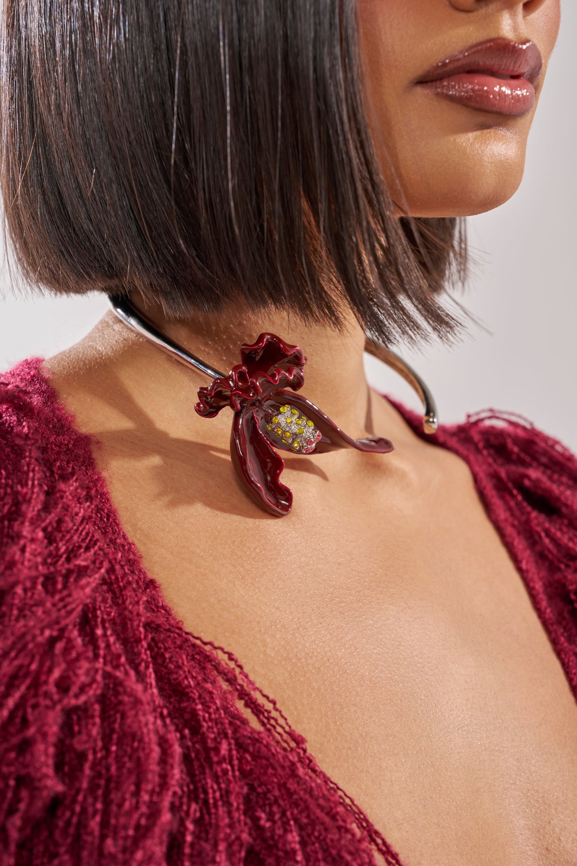 A woman with a sleek bob wears a textured deep red top and the FULL BLOOM NECKLACE, featuring a metallic statement design with a glossy red enamel flower and yellow accents. 0