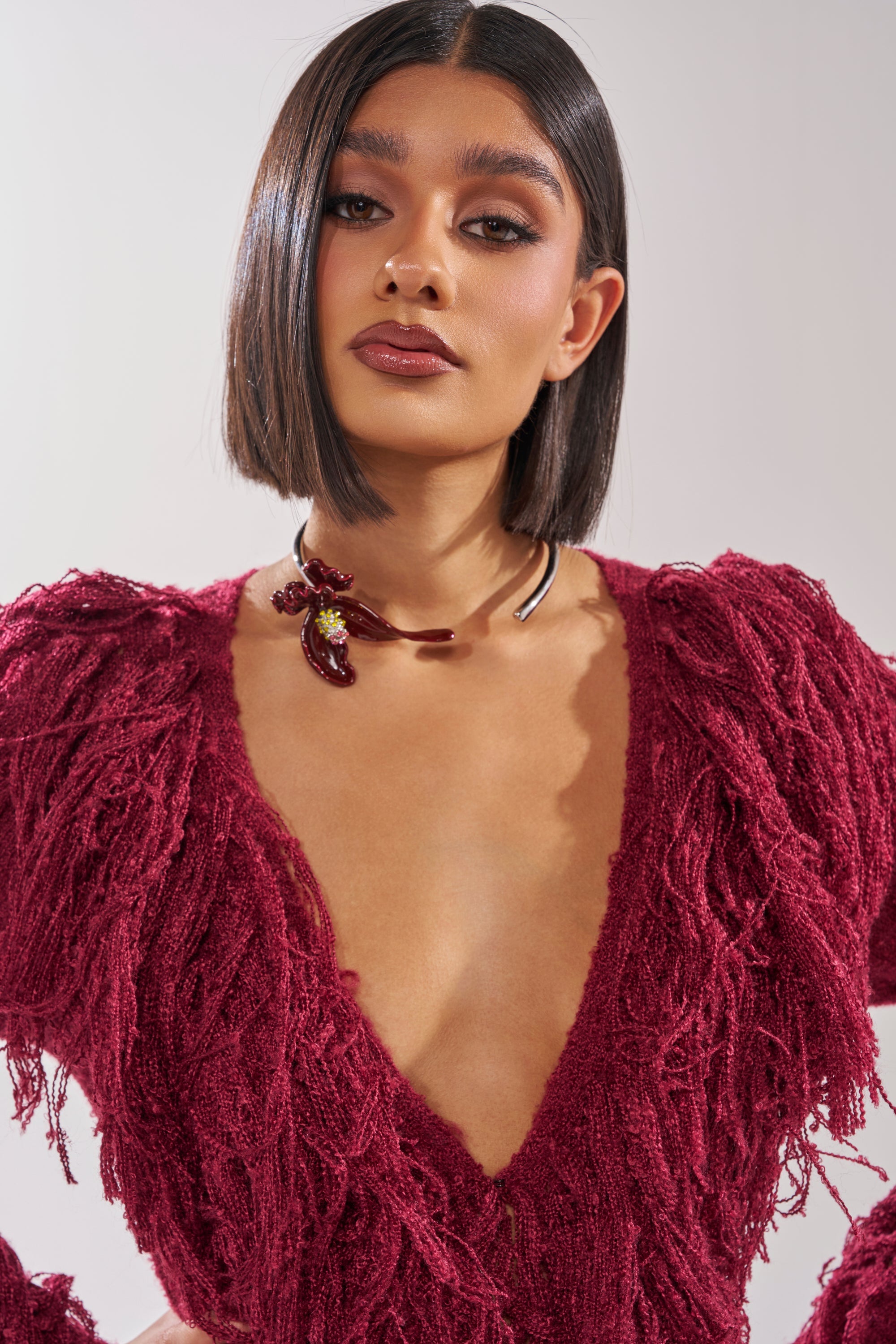 A person with a sleek bob hairstyle wears a deep red, textured V-neck top and the FULL BLOOM NECKLACE, featuring floral details, posing confidently against a plain light background. 1
