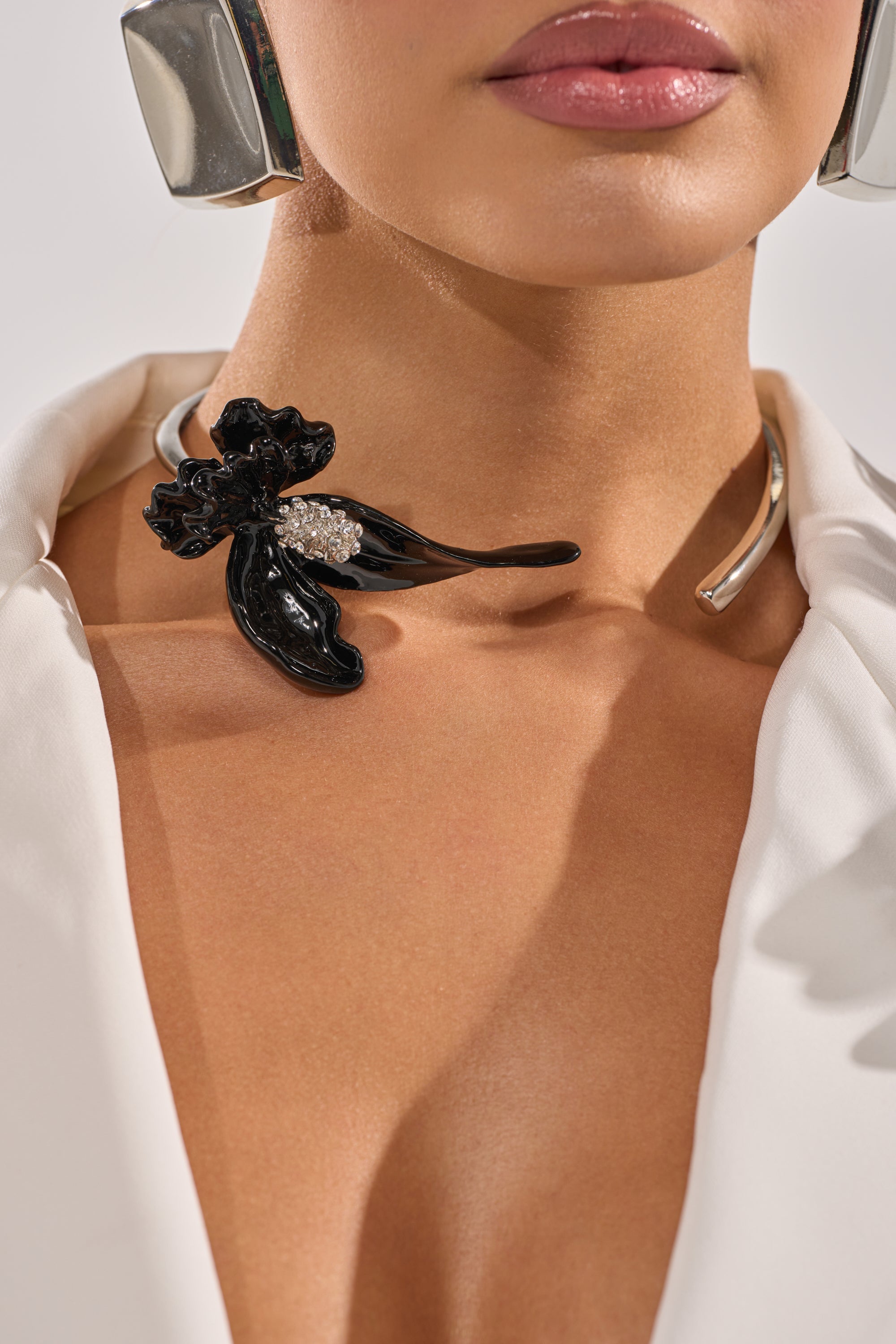 A close-up of a person wearing the FULL BLOOM NECKLACE, a silver-tone collar with a black floral accent and rhinestone details, styled with a white blazer and large silver earrings. Only their lips and part of the jaw are visible. 0