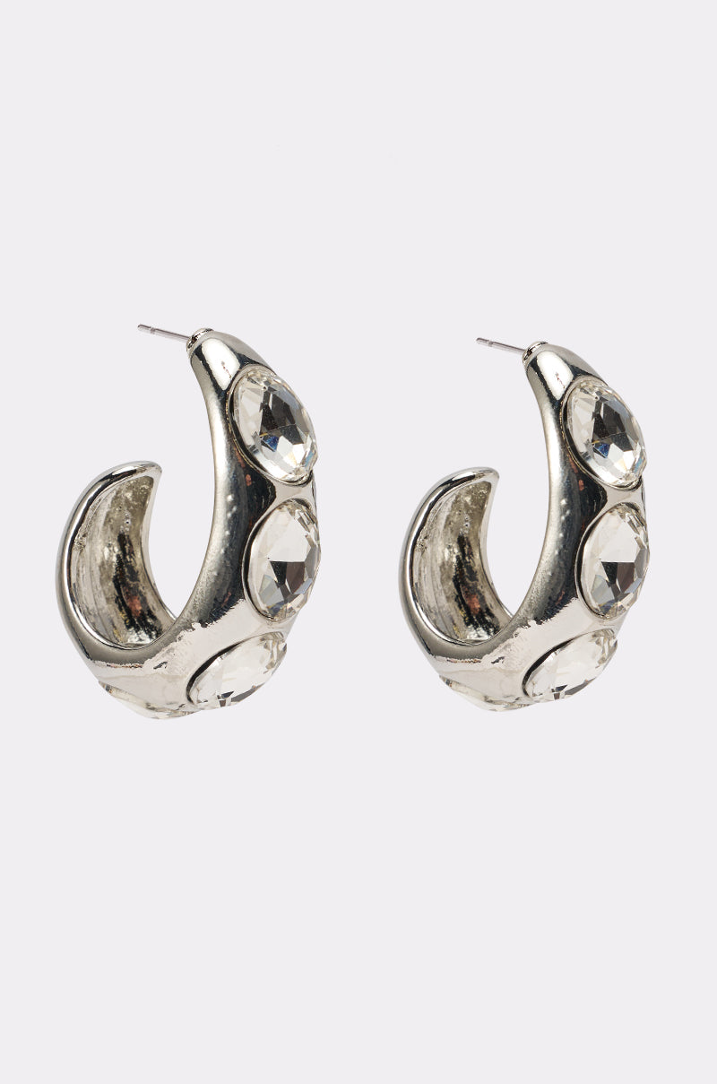 The KELYN EARRING features chunky silver hoops adorned with three large faceted clear gemstones, creating dazzling statement earrings shown against a plain white background. 0