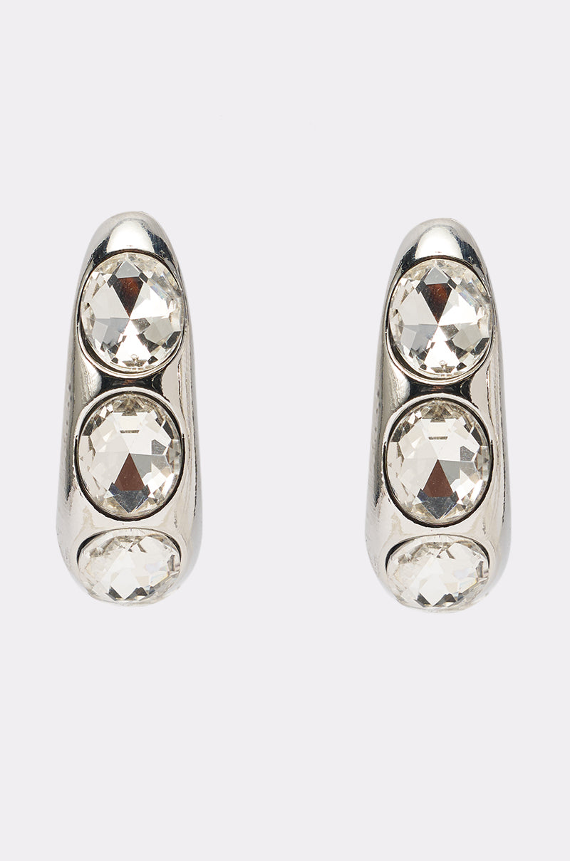 The KELYN EARRING is a pair of silver-toned statement earrings, each with three round clear gemstones set vertically in a smooth, rounded metal frame, shown on a light background. 3