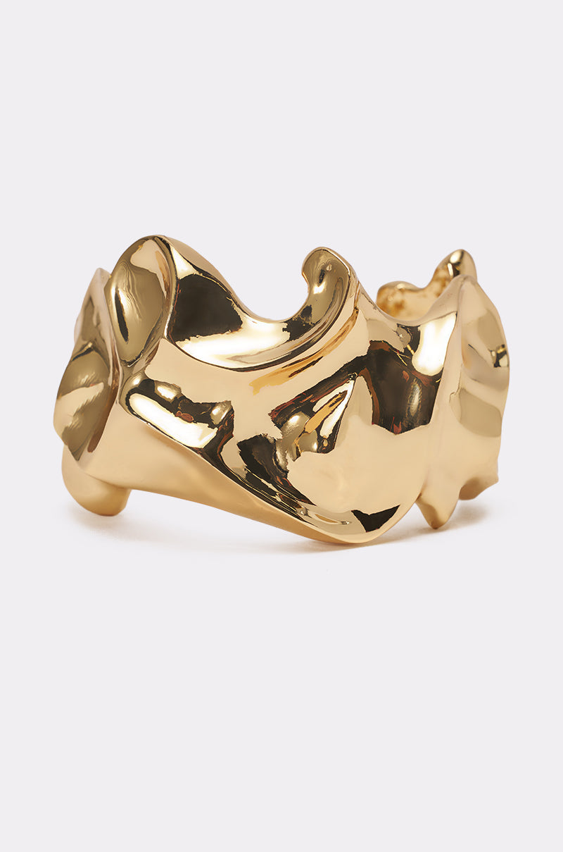 The MOLTEN DREAMS BRACELET IN GOLD features a shiny, abstract wavy design and is displayed against a plain light background. 0