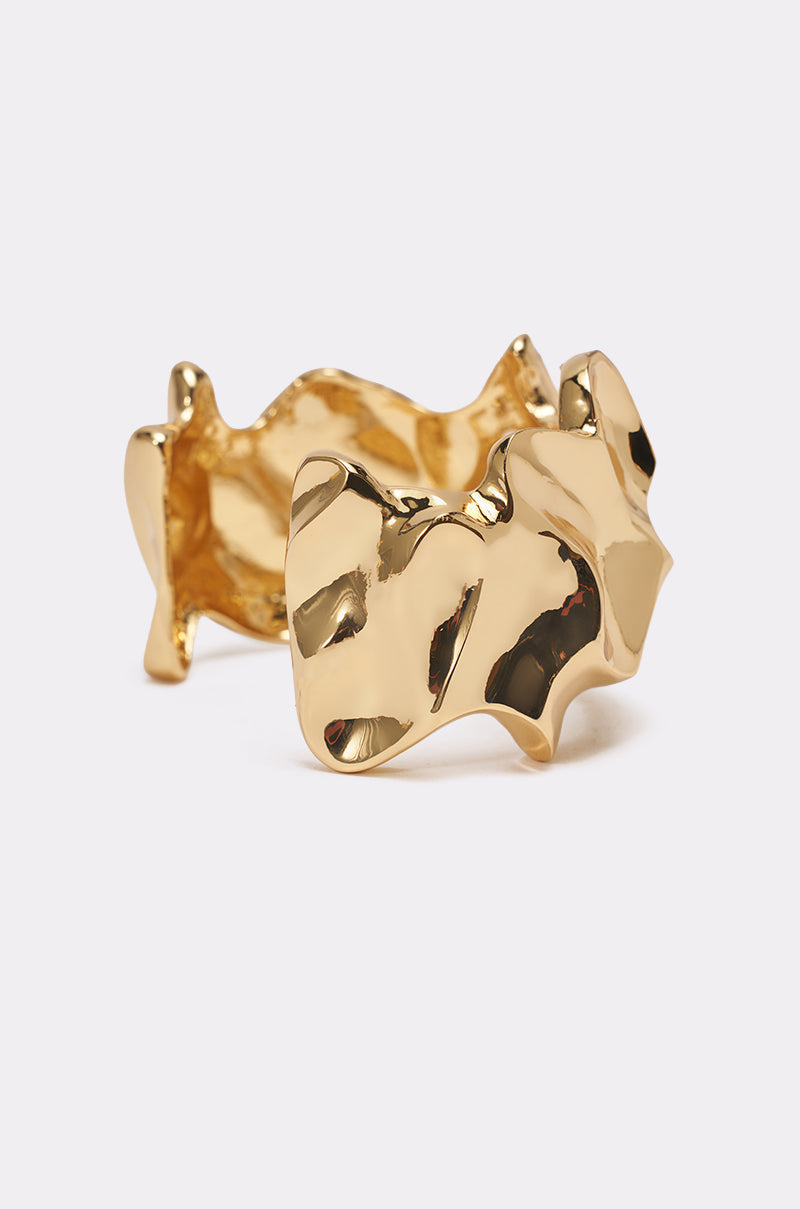 A close-up of the MOLTEN DREAMS BRACELET IN GOLD showcases its wide, abstract, wavy, and textured surface—resembling hammered gold—for a bold statement look against a plain white background. 2