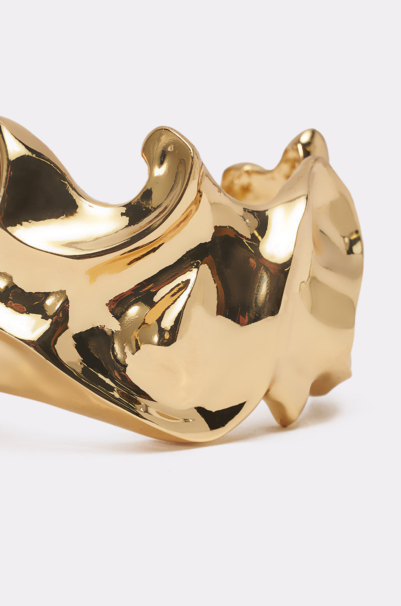 A close-up of the MOLTEN DREAMS BRACELET IN GOLD, featuring smooth, curved surfaces and reflective highlights that evoke an abstract gold sculpture, set against a plain light background. 3