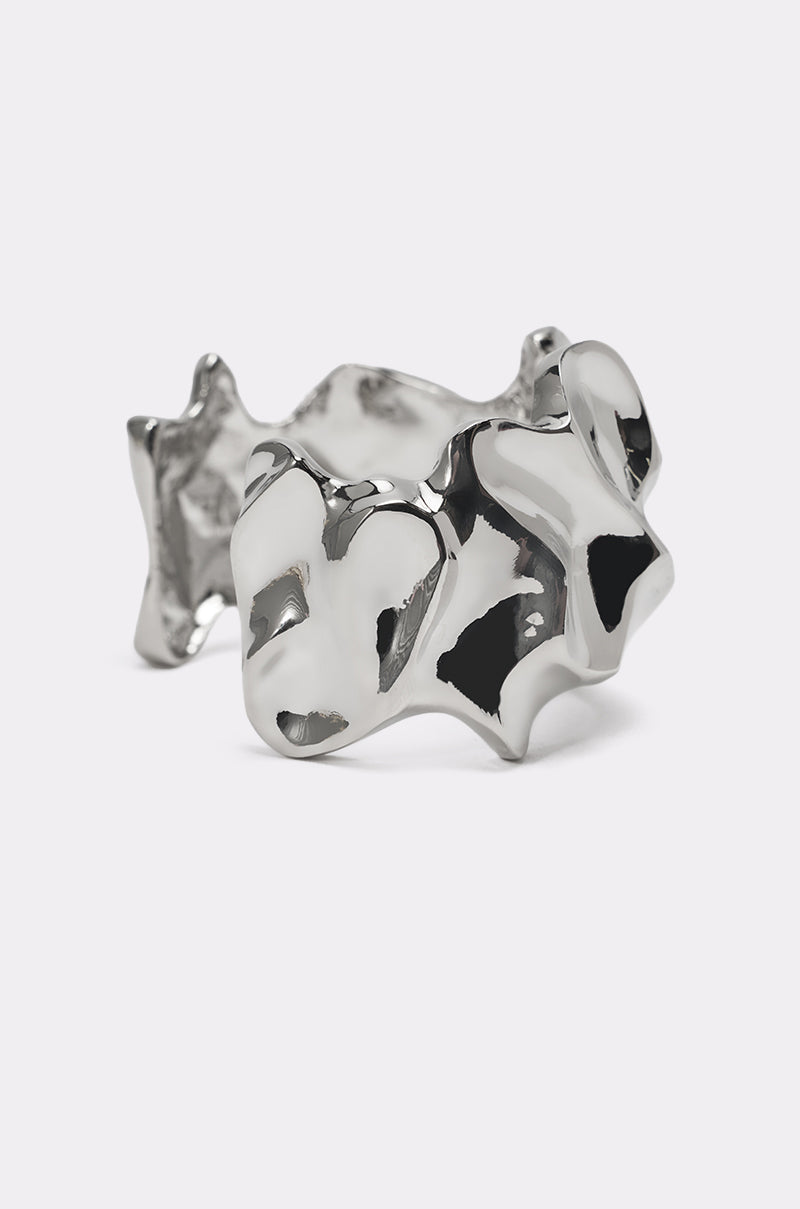 The MOLTEN DREAMS BRACELET features a chunky, irregular silver design with a glossy, reflective finish, embodying bold minimalist style and showcased on a light gray background. 2
