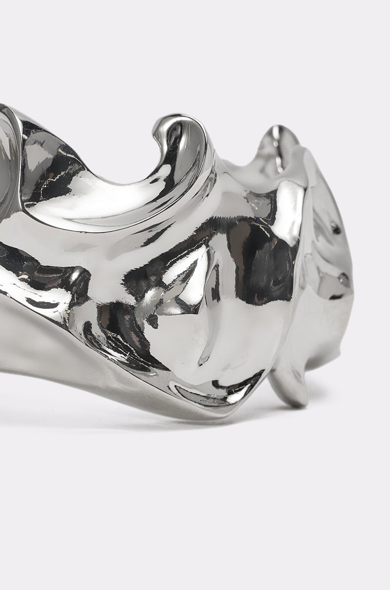 A close-up of the MOLTEN DREAMS BRACELET reveals its highly polished silver surface and abstract, flowing curves reflecting light. This sculptural cuff stands out brilliantly against a plain, light-colored background. 3