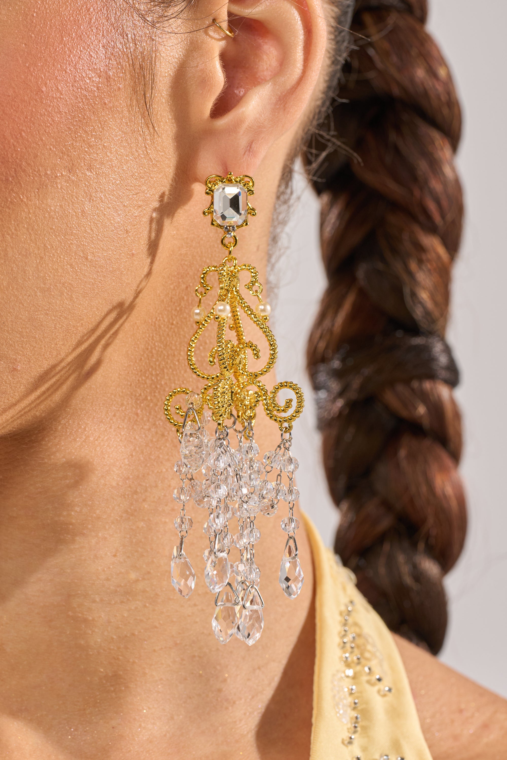 Close-up of a person wearing the SEE MY LIGHT EARRING—ornate gold chandelier earrings with clear crystal beads and delicate fringe. The ear and braided brown hair are visible, highlighting these striking statement earrings. 1
