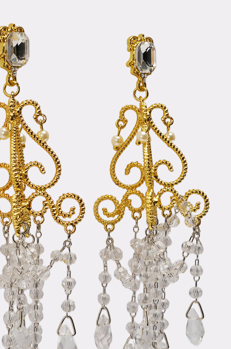Close-up of SEE MY LIGHT EARRING—ornate gold chandelier earrings with intricate scrollwork, small pearls, and dangling clear crystal beads on a light background, perfect as a statement piece for an elegant look. 3
