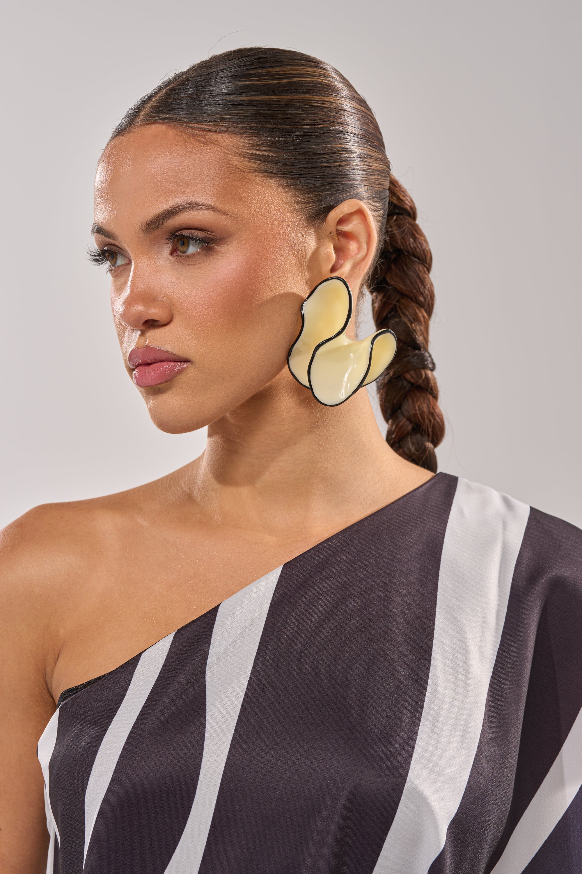 A woman with a sleek, braided ponytail wears the bold yellow ITS MY WORLD EARRING and a black-and-white, one-shoulder striped top, gazing to the side against a neutral background. 1
