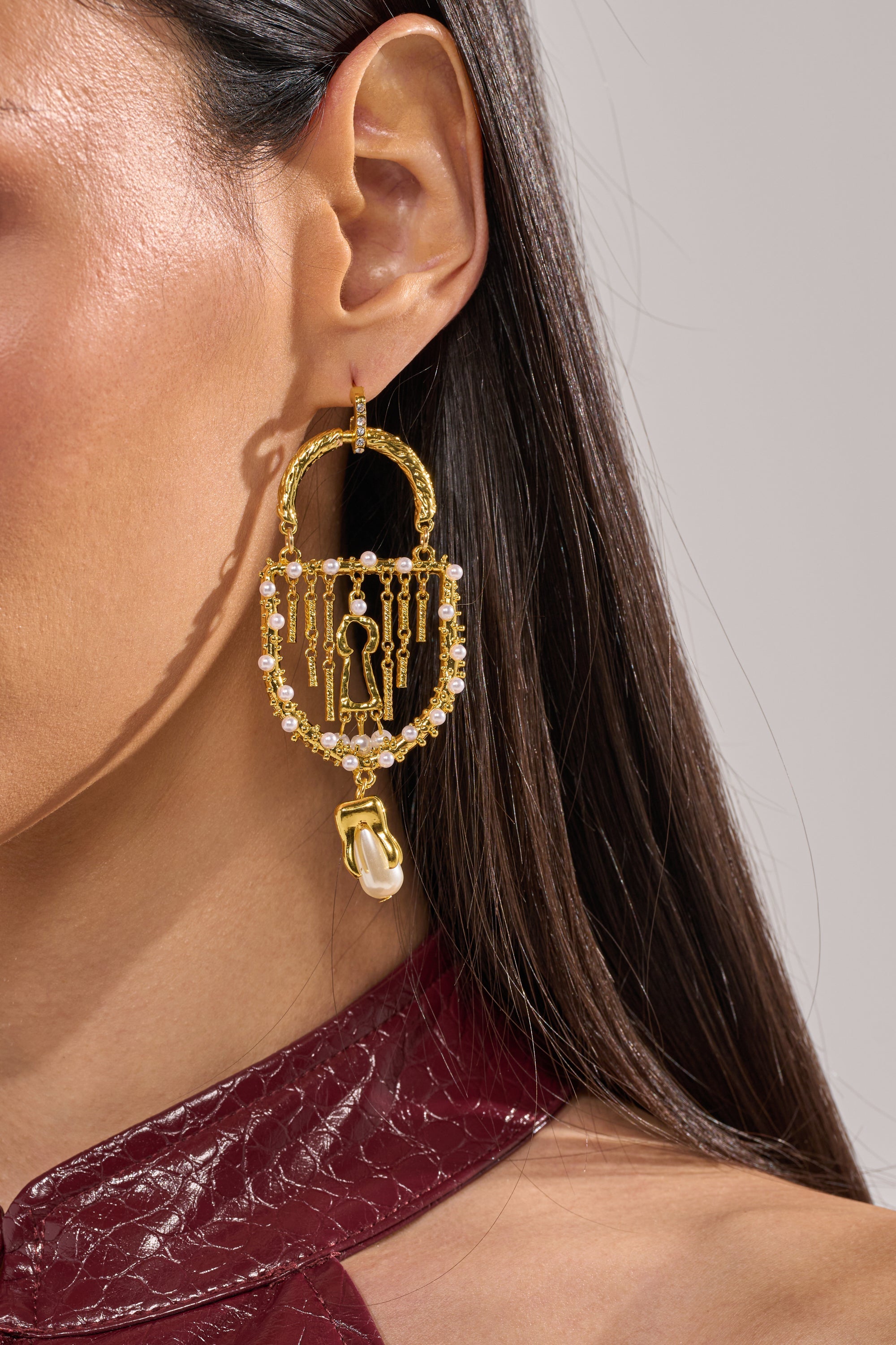 Close-up of a woman wearing the UNLOCK ME EARRING—gold statement earrings shaped like padlocks with dangling key and pearl charms—paired with a burgundy textured top against a neutral background. 1