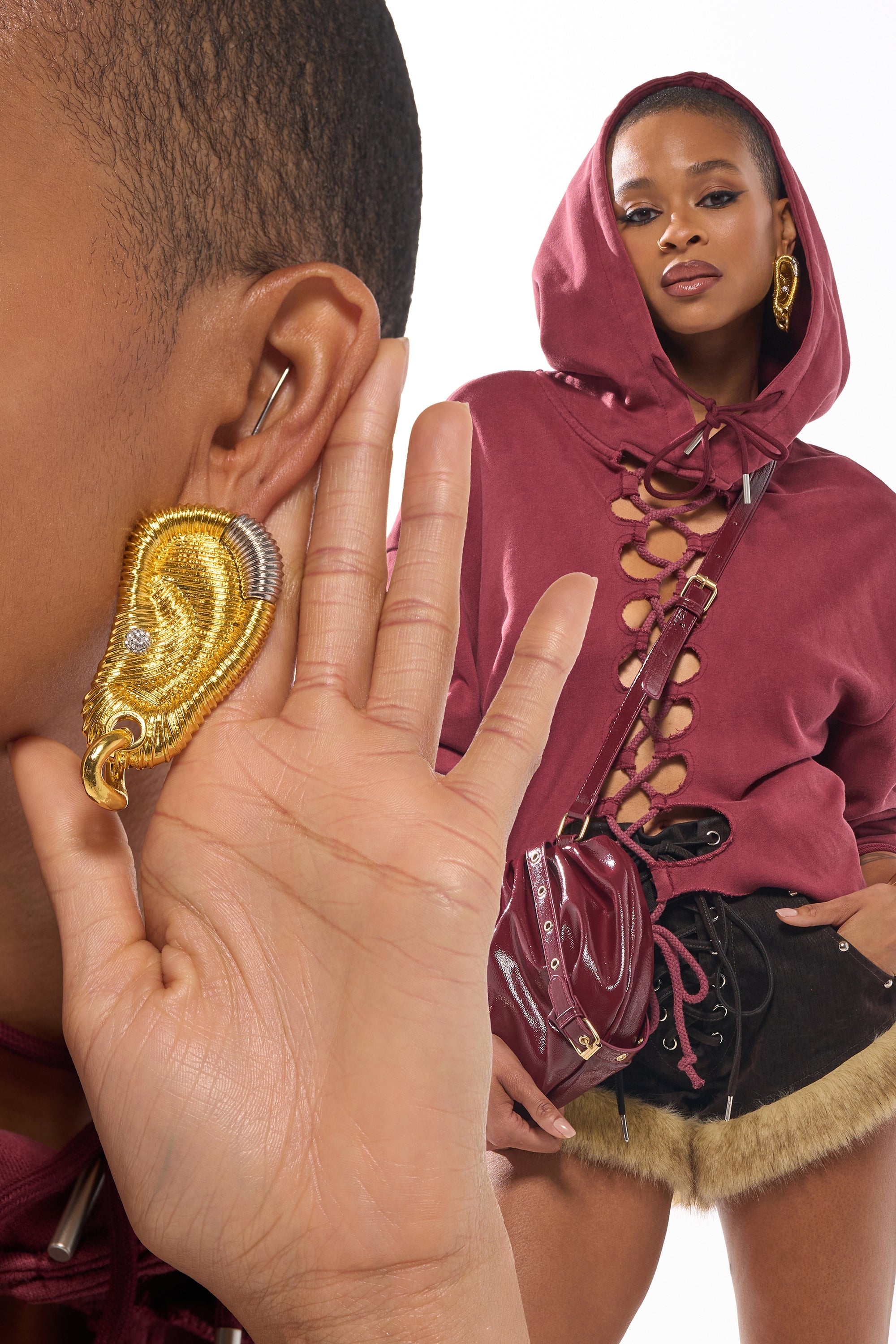A woman in a maroon lace-up hoodie and black fur-trim shorts highlights her sculptural HEARD U EARRING while also wearing gold statement earrings and carrying a maroon crossbody bag. 0