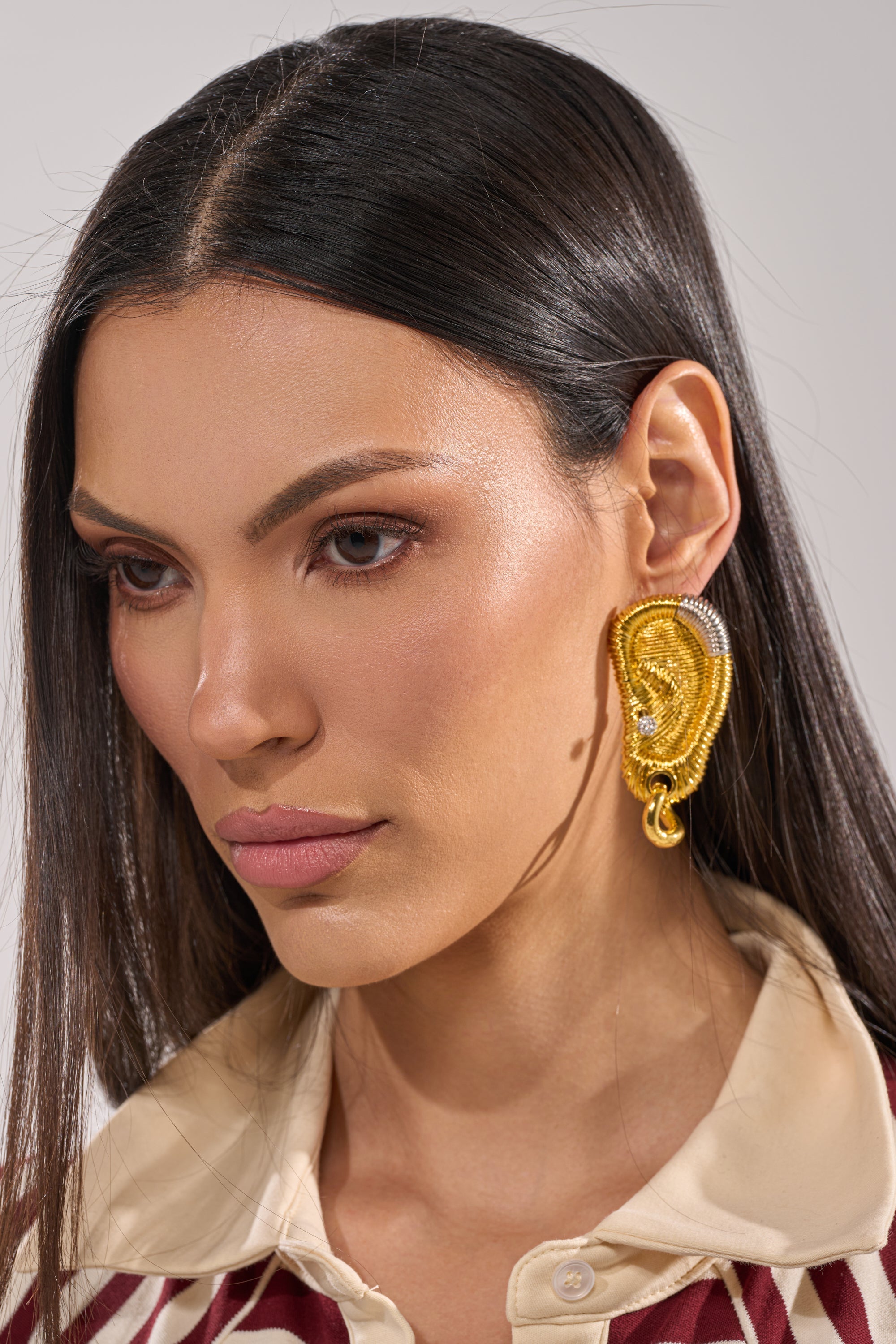 A woman with straight dark hair wears sculptural, gold HEARD U EARRINGS and a beige collared shirt with a patterned design. She has natural makeup and gazes slightly downward with a neutral expression. 1