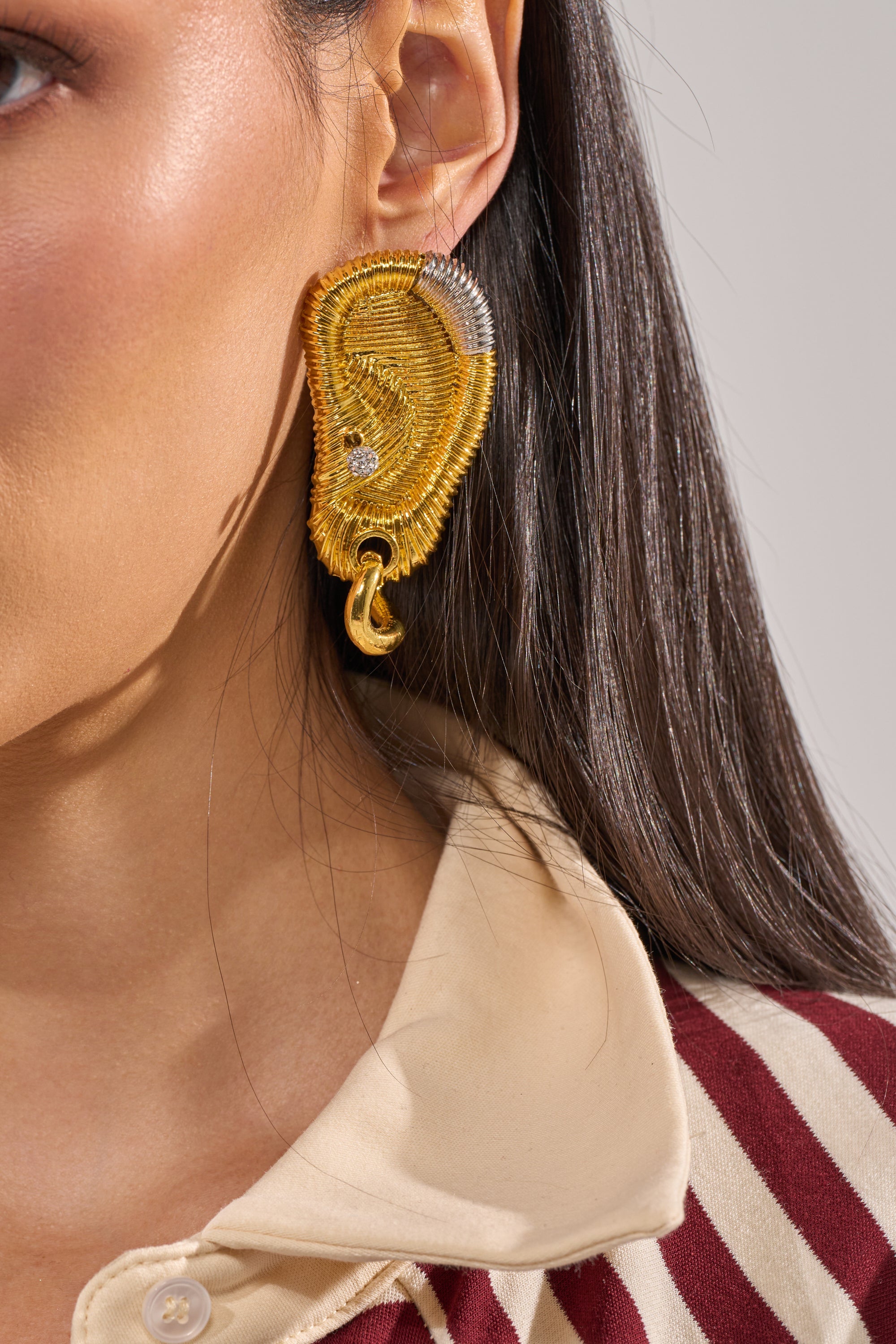 Close-up of a person with long dark hair in a cream and maroon striped shirt wearing the HEARD U EARRING—sculptural gold statement earrings shaped like an ear, accented with a small gem. 5