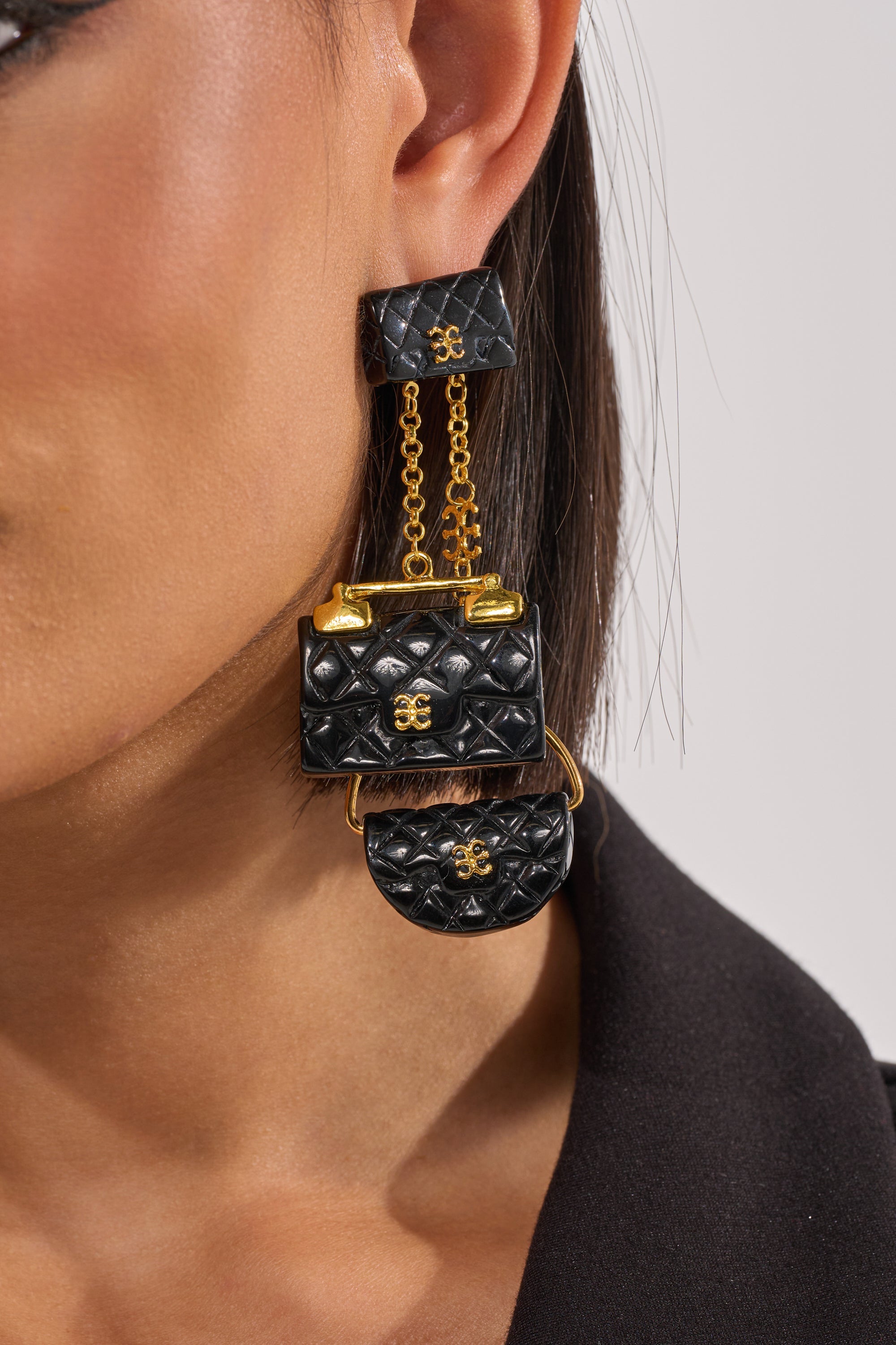 Close-up of a woman's ear wearing the GET ME A BAG EARRING, designed as miniature black quilted handbags with gold hardware and chain details. She's in a black top, shown against a neutral background. 1