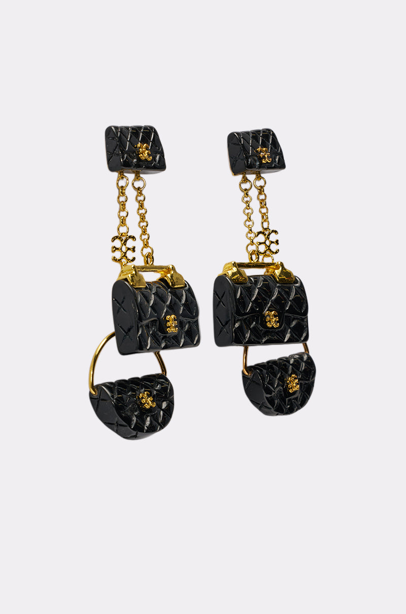 The GET ME A BAG EARRING is a pair of black and gold quilted purse-shaped earrings with gold chain details and small gold accents—perfect statement pieces, displayed on a plain light grey background. 2