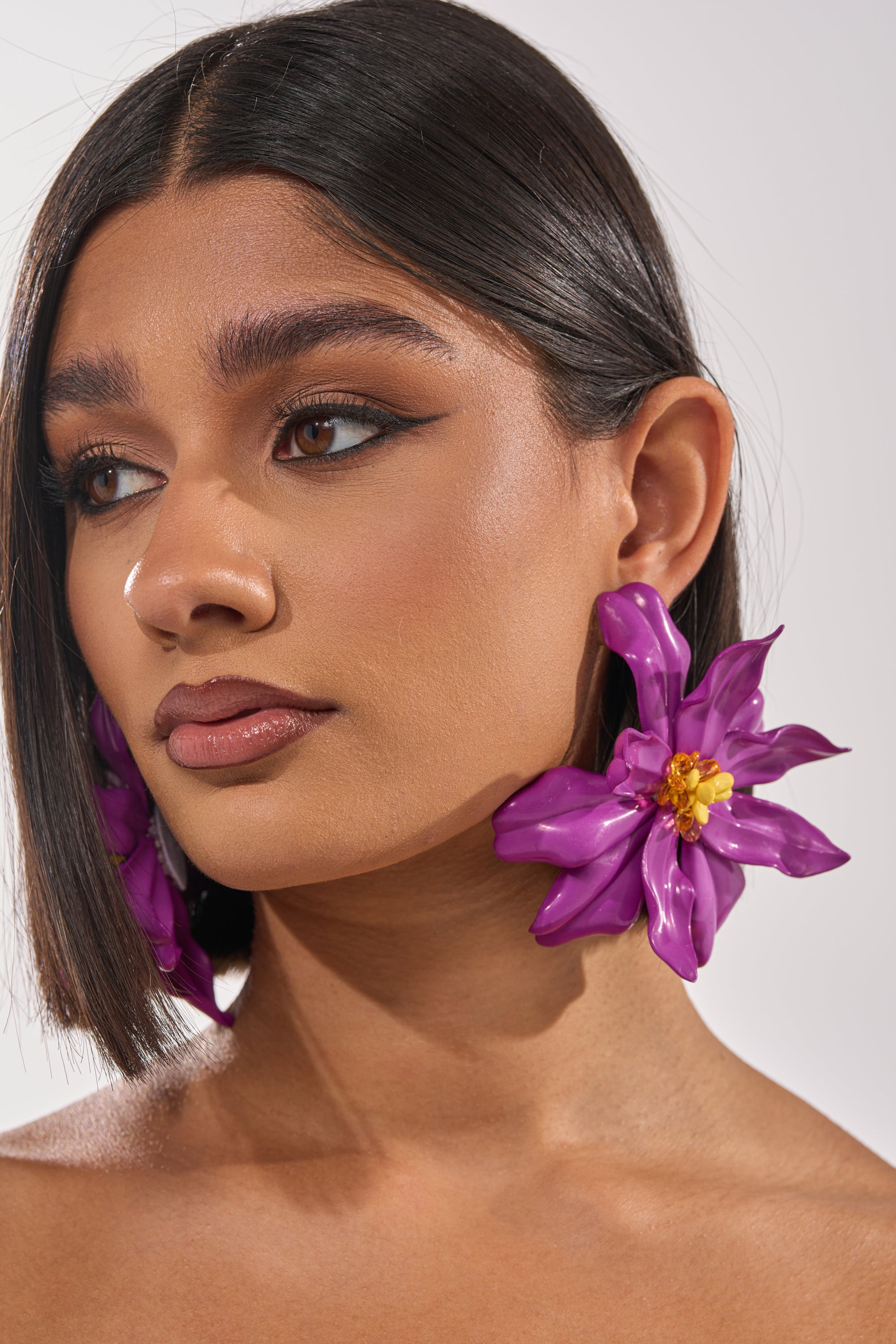 A woman with straight dark hair and winged eyeliner wears the IN THE GARDEN EARRING, featuring bold purple floral accents. She gazes calmly to the side against a plain background. 1