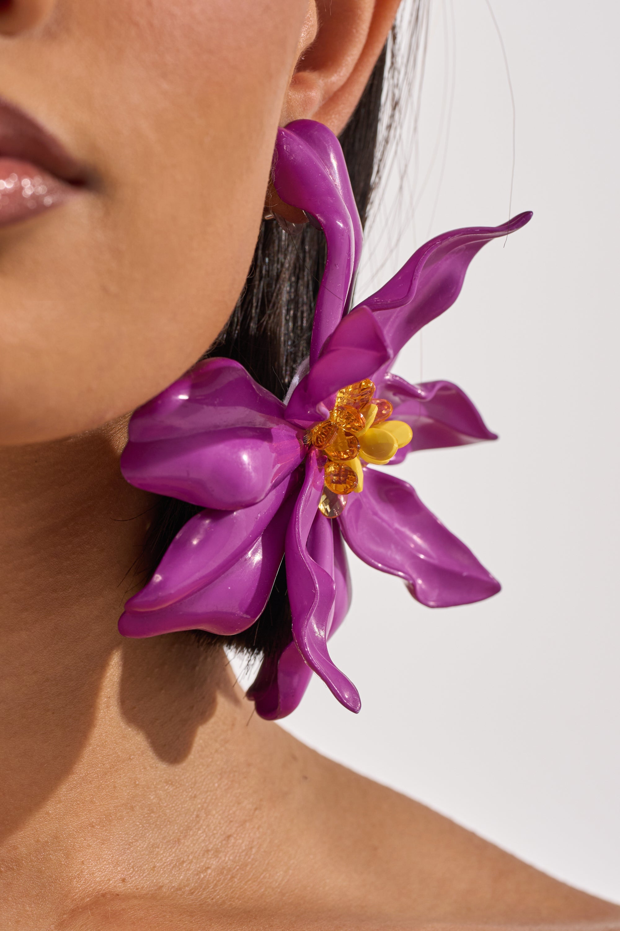 A close-up of a person wearing IN THE GARDEN EARRING—oversized, orchid-shaped earrings in vibrant purple and yellow. These tropical-inspired earrings pop against their skin and short, dark hair. 4