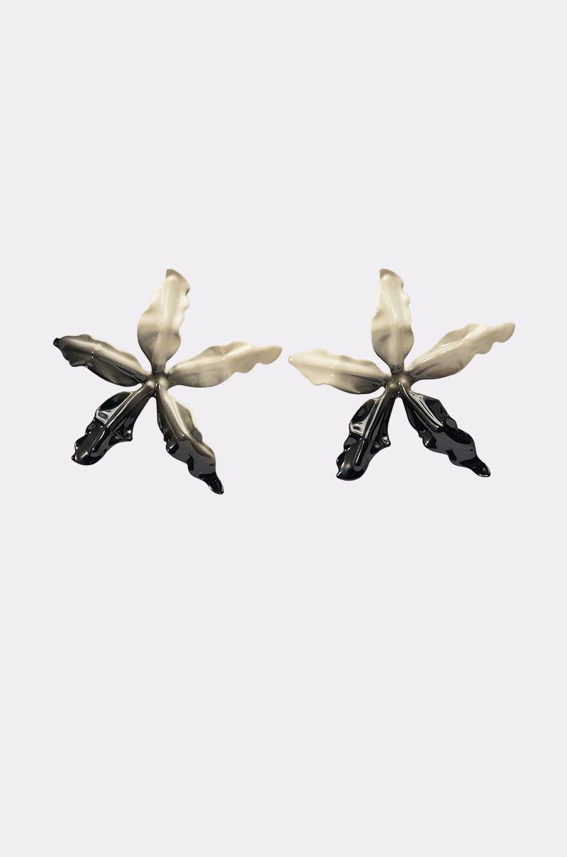 The TAKE A TRIP EARRING features oversized five-petaled flower shapes in metallic silver and black, displayed on a plain light gray background. 0