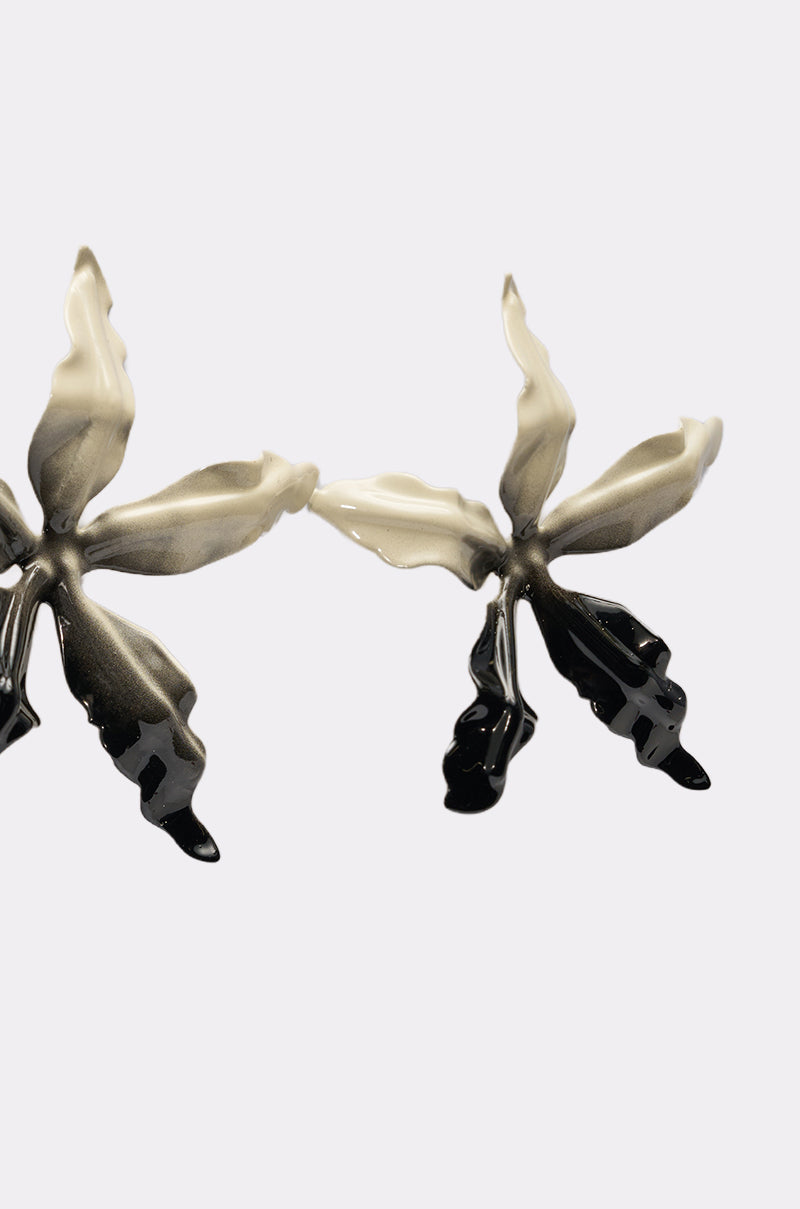 The TAKE A TRIP EARRING features two metallic, oversized statement earrings in an orchid shape with a silver-to-black gradient, shown against a plain white background. 3