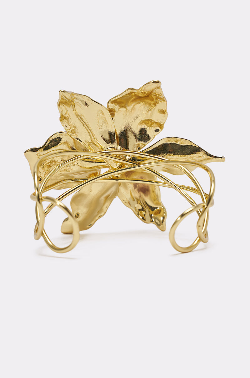 The BY THE SHORE BRACELET is a gold-tone cuff with a bold, textured five-petal flower at the center and swirling, vine-inspired bands wrapping elegantly around the wrist. 3