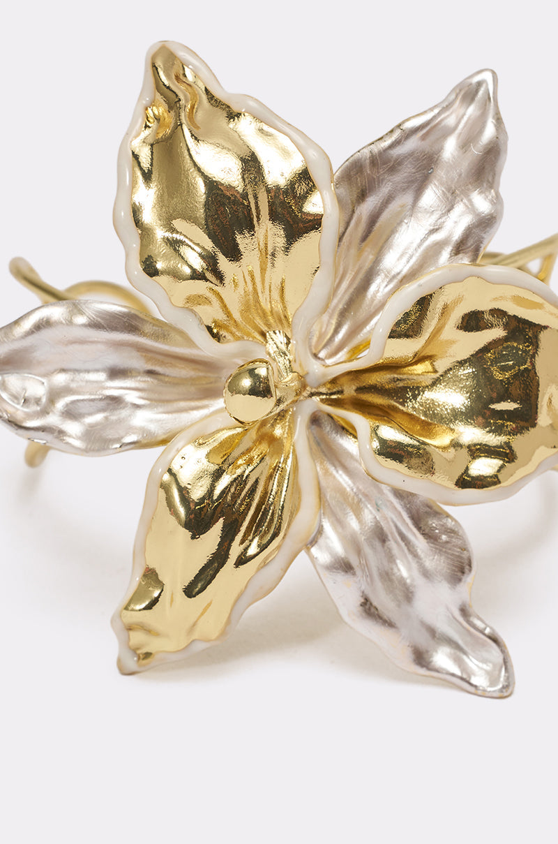 A close-up of the BY THE SHORE BRACELET, a sculptural flower jewelry piece with two gold petals and three silver petals, featuring a glossy, reflective finish on a light background. 4