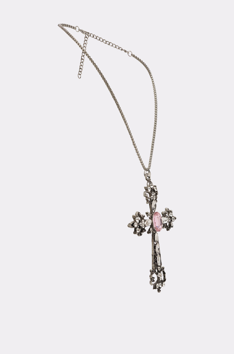 The LEXI NECKLACE is a silver cross necklace with an ornate rhinestone cross pendant, sparkling clear stones, and a large pink gem at the center, displayed on a plain white background. 3