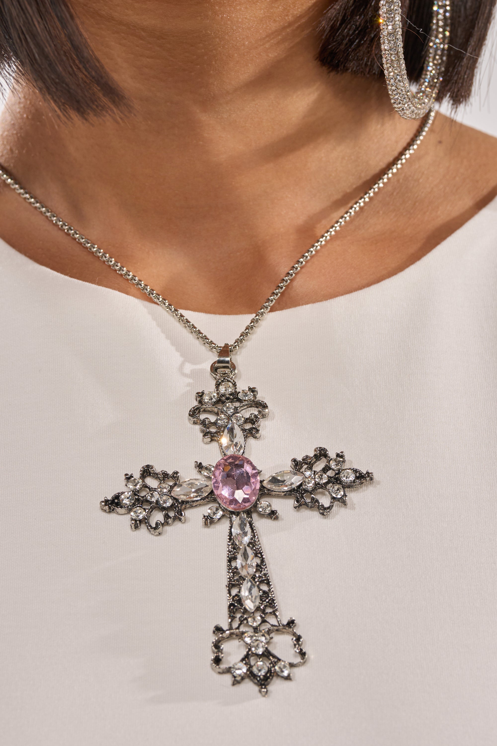 A woman wears a white top, large hoop earrings, and the LEXI NECKLACE—a silver cross with an ornate pendant highlighted by a large pink gemstone in the center and smaller clear gems. 5