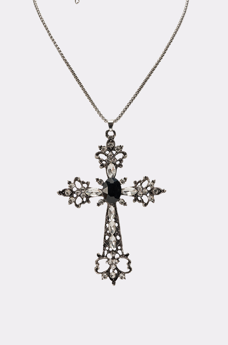 The KIM NECKLACE is a silver-finish cross necklace with an ornate filigree design, featuring small clear rhinestones and a striking large black gemstone at the center, displayed on a plain white background. 3
