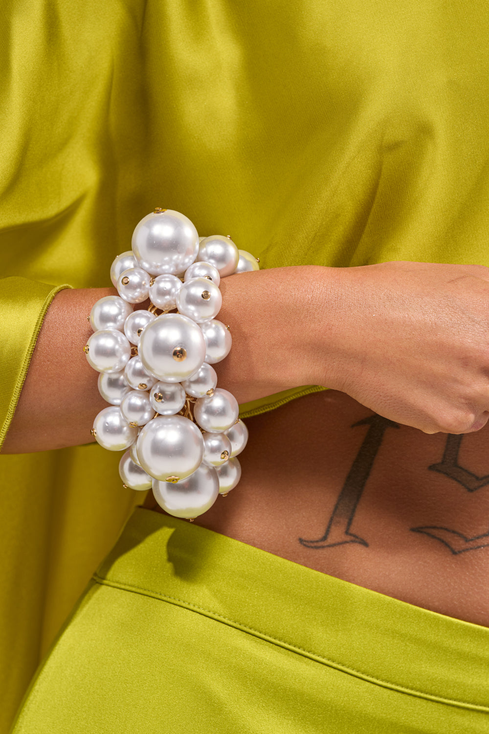 Wearing a shiny lime-green outfit, a person flaunts the FEELIN BUBBLY BRACELET—a chunky cuff with large and small white pearls. Their midriff, showing part of a tattoo with the letters "L" and "I," is visible.