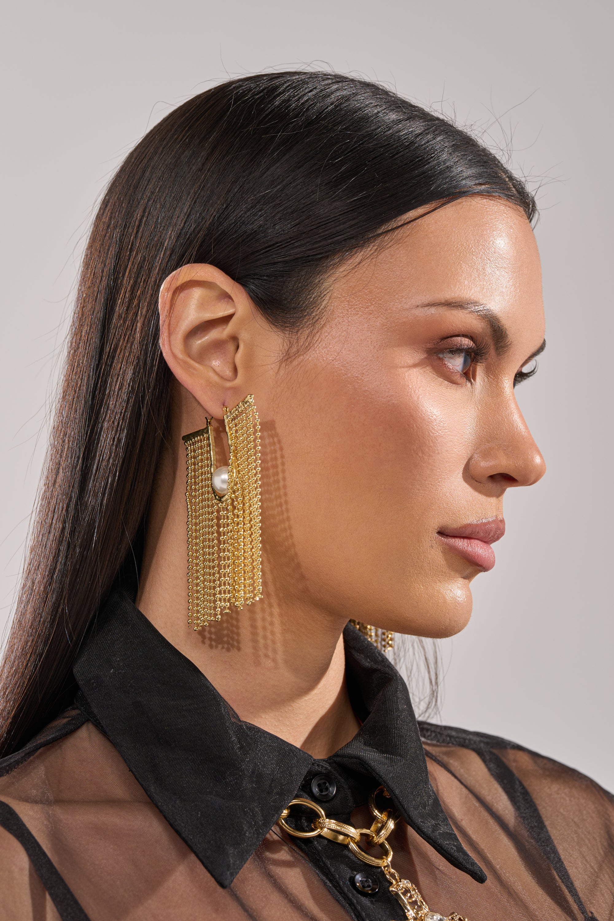 A woman with straight dark hair wears the U DONT GET IT EARRING—pearl tassel earrings with gold-tone chains and dangling strands—paired with a sheer black collared top, posing in profile against a neutral background. 4