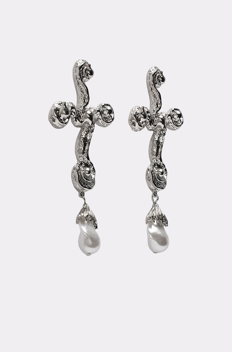The CROSS ME EARRING features a pair of silver cross-shaped earrings with an organic textured design and dangling white pearl drops, showcased against a simple light background for a striking statement look. 2