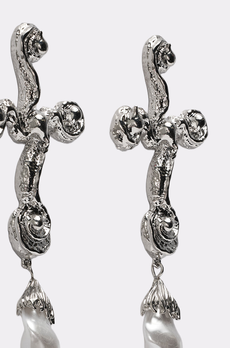 The CROSS ME EARRING features two silver-tone, abstract cross-shaped earrings with textured, rounded forms and a dangling teardrop-shaped white pearl-like bead. Shown against a plain light background. 3