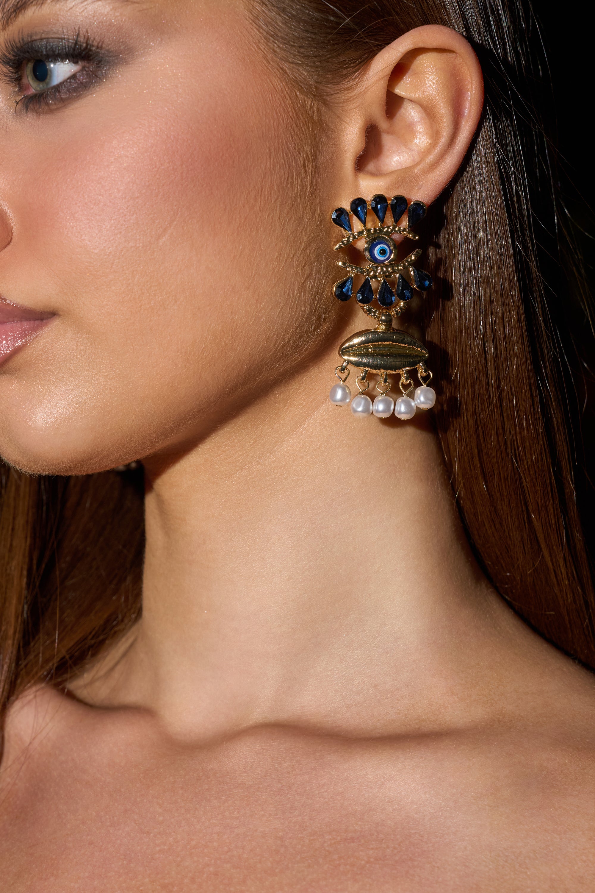 Close-up of a woman wearing the TWO FACED EARRING—gold eye earrings with blue and gold details, hanging white pearls, and bold design. Her straight brown hair is tucked back, highlighting these striking festival-ready statement earrings.