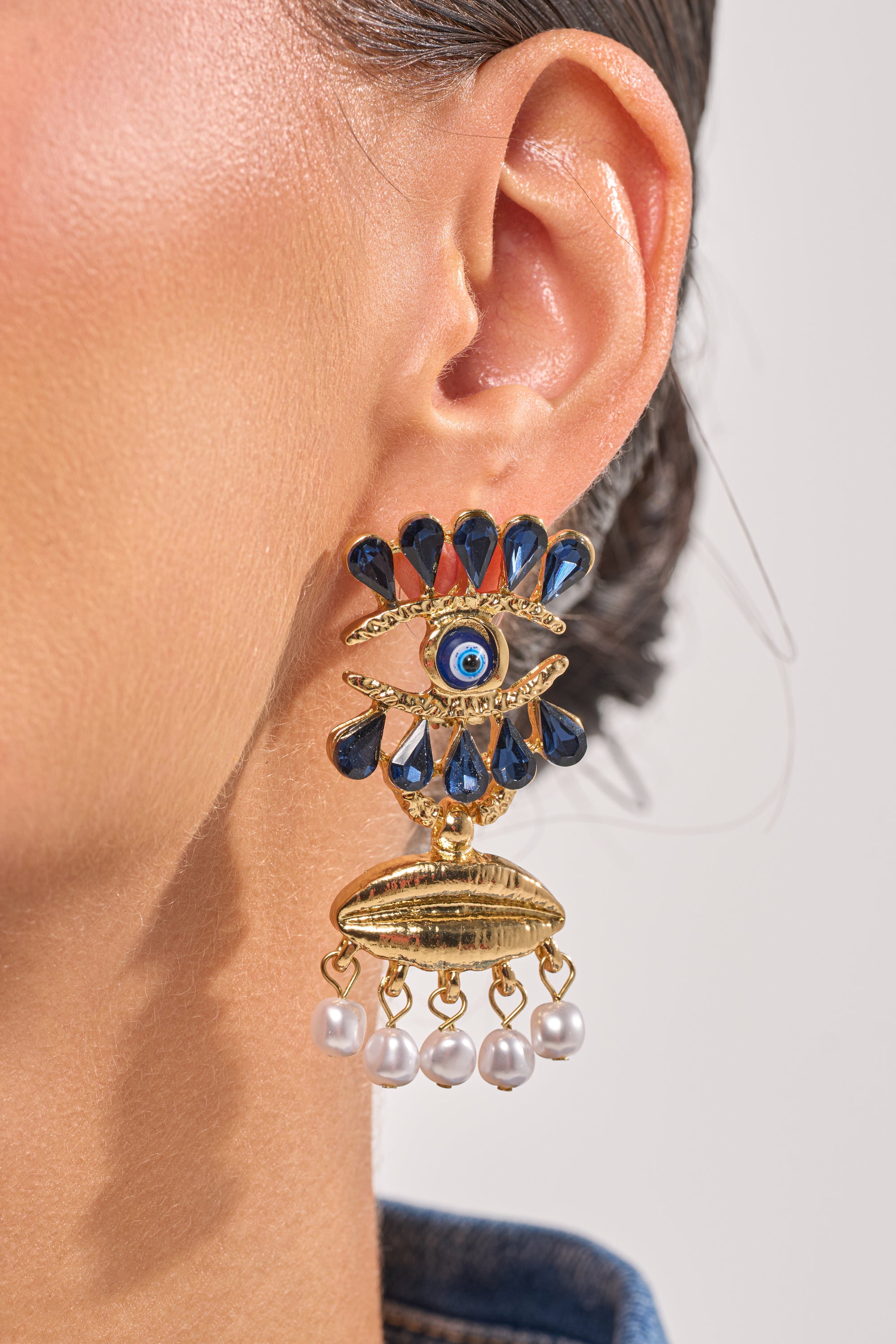 Close-up of a woman's ear wearing the TWO FACED EARRING—bold gold eye earrings with blue evil eye design, teardrop stones, gold accents, and dangling pearls—perfect statement accessories for festivals. 1