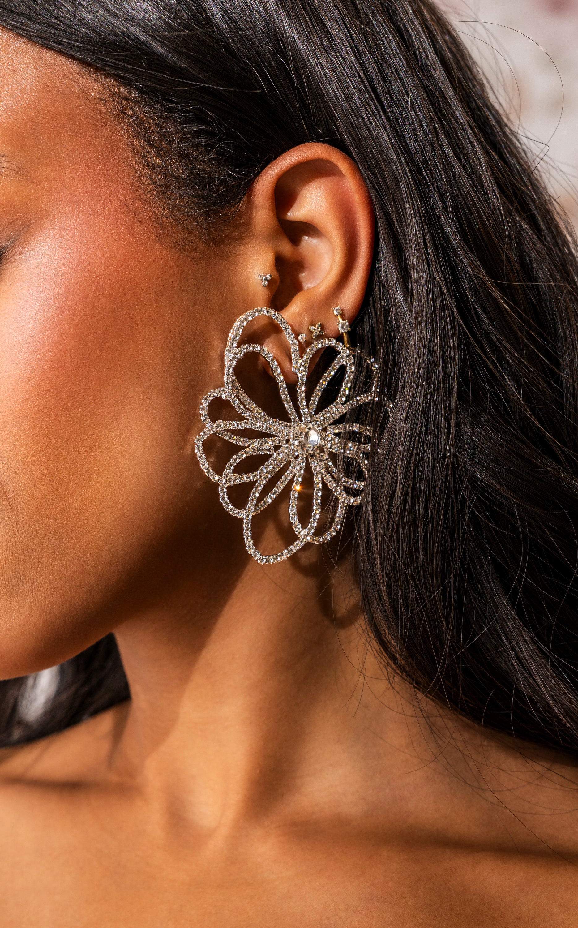 A close-up of a woman with dark, wavy hair wearing the IM OUTSIDE EARRING—a large, sparkling rhinestone floral statement piece with crystal details—in her left ear, styled with a few small studs above it. 0