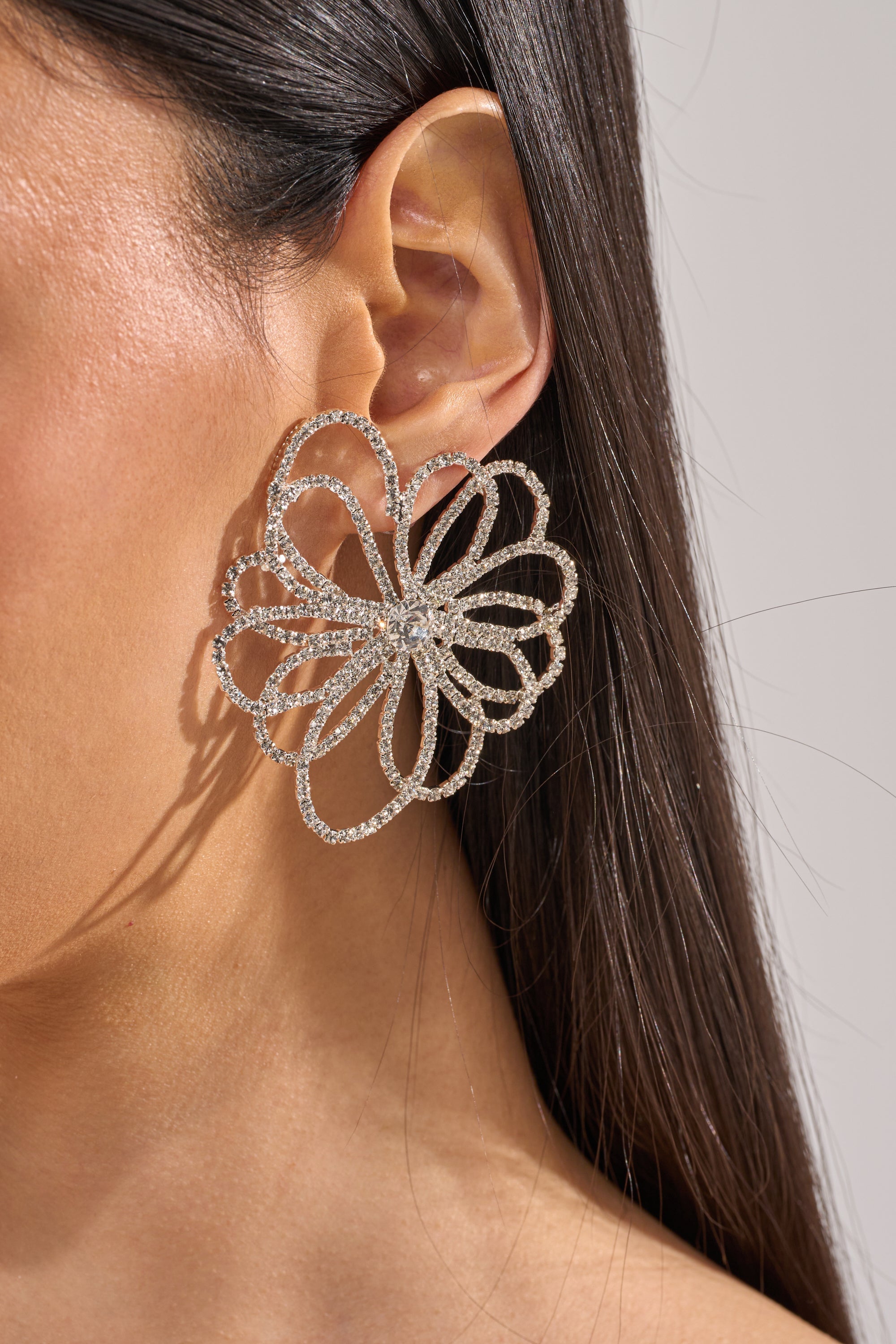 A close-up of a woman's ear wearing the IM OUTSIDE EARRING—large, sparkling rhinestone floral earrings in silver. Her straight, dark hair is tucked to reveal these stunning flower-shaped statement earrings. 2