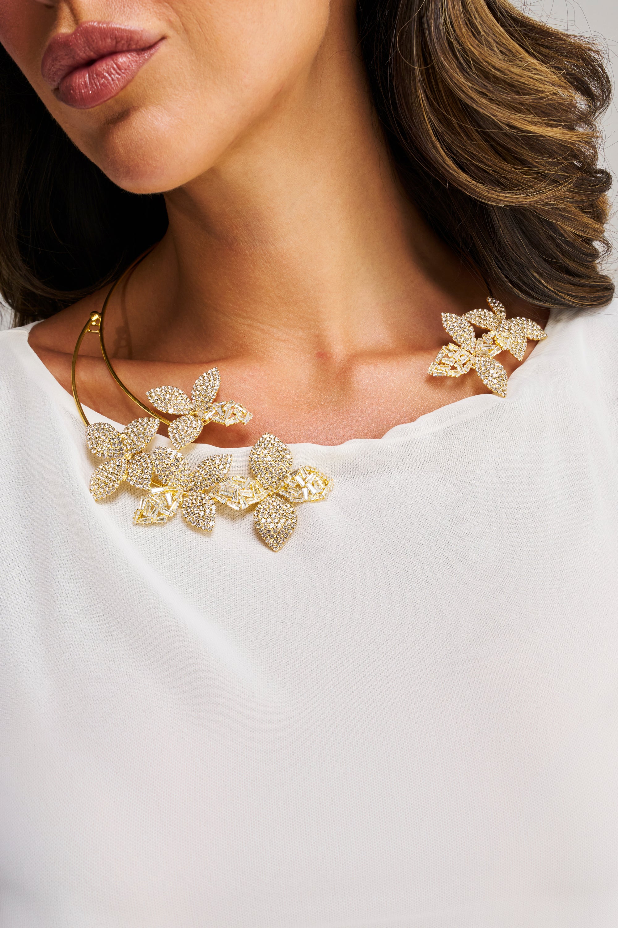 A woman wears the IN BLOOM NECKLACE, a gold-tone collar adorned with sparkling crystals, paired with a white blouse. The image is cropped to highlight her lips, neck, and part of her shoulder.