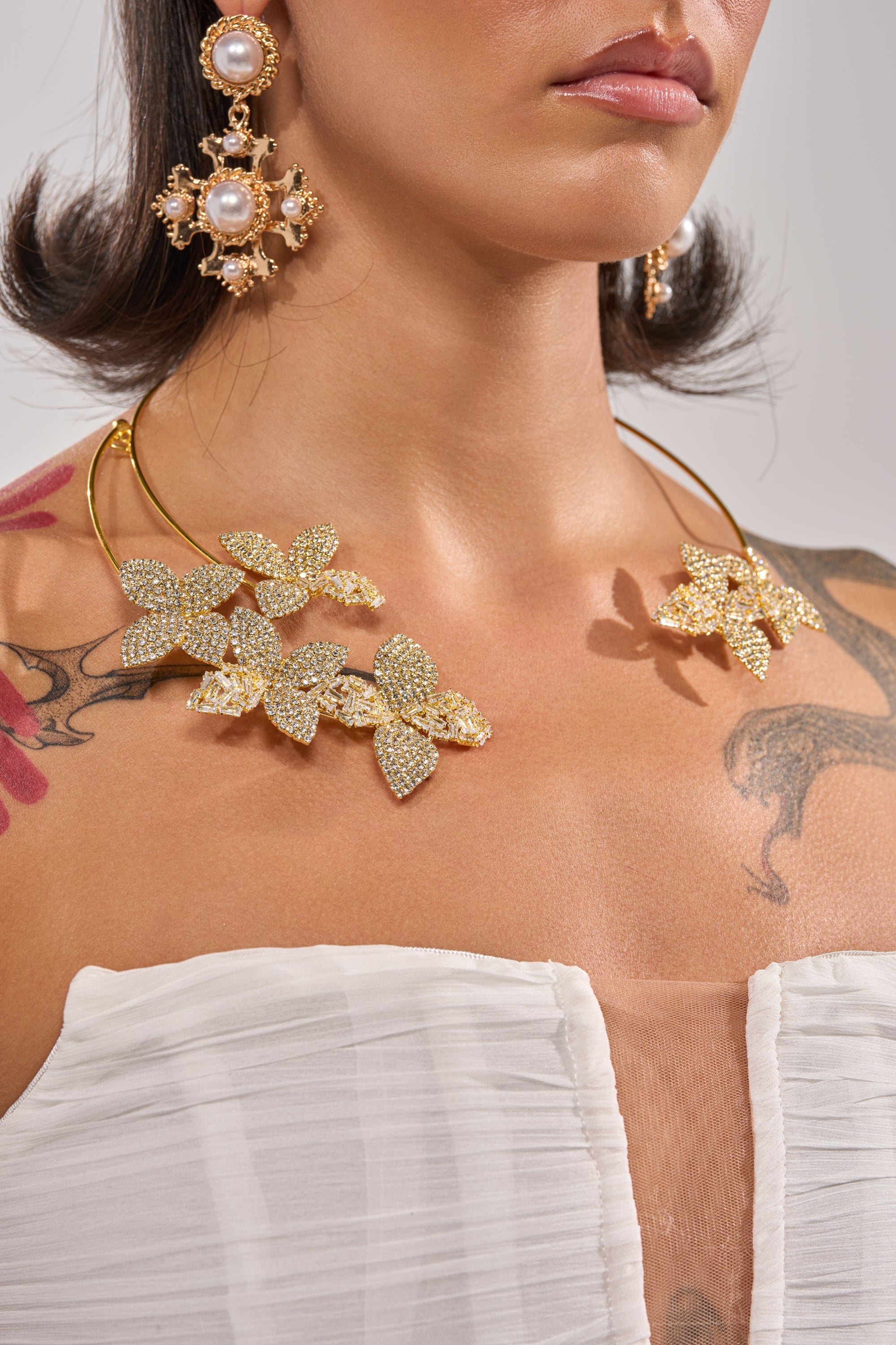A woman wears a white strapless top, large pearl drop earrings, and the IN BLOOM NECKLACE—a rhinestone floral statement piece. Part of her face, neck, and tattooed upper chest are visible. 1