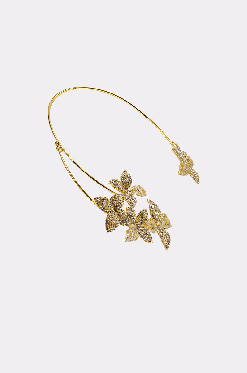 The IN BLOOM NECKLACE is a gold-tone open-neck collar featuring two branches, each ending in sparkling floral rhinestone designs, shown on a light background. 2