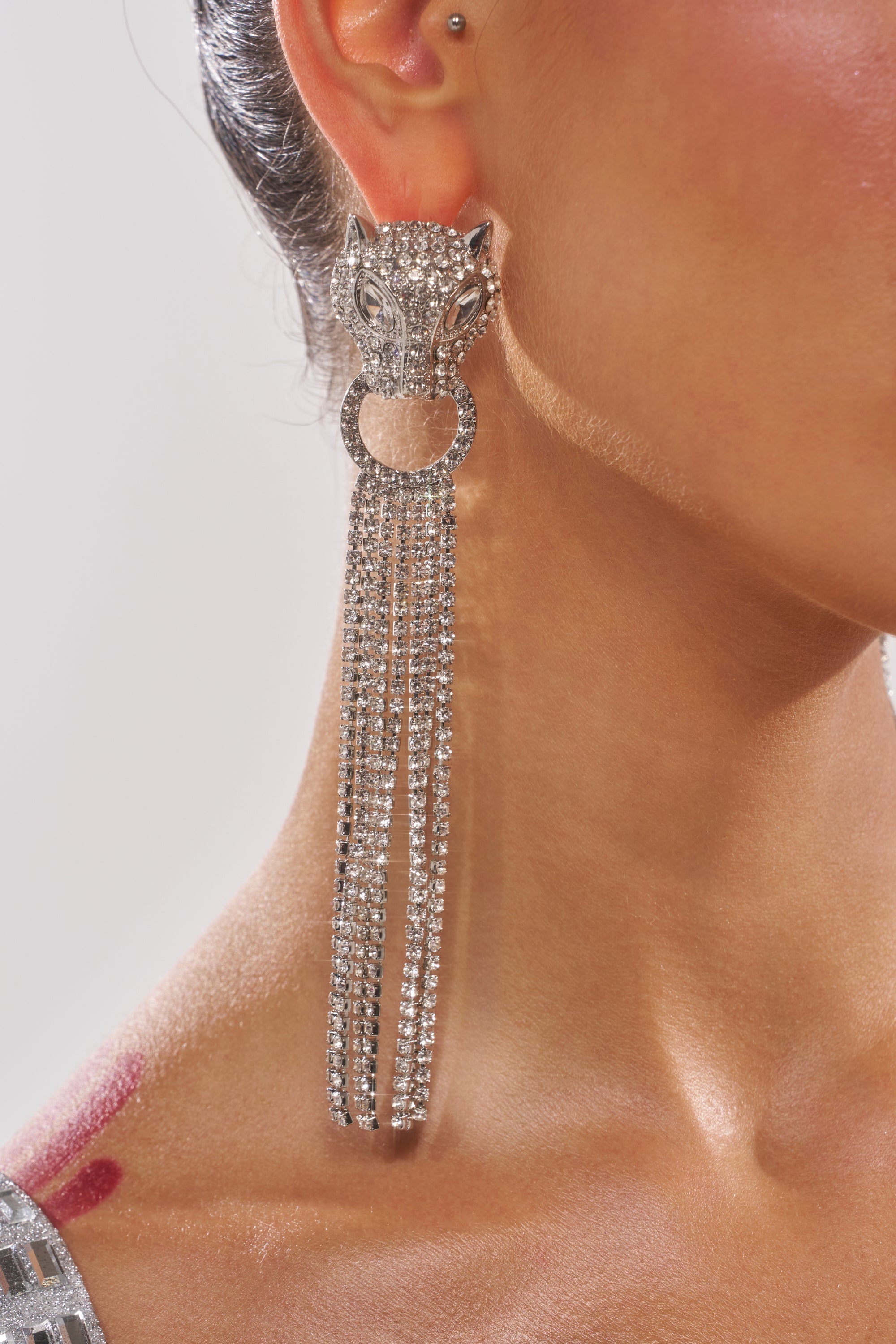 A close-up of a woman's ear wearing the AT THE GALA EARRING, designed as a leopard's head with a ring in its mouth and rhinestone fringe. A visible burgundy mark appears on the skin near her shoulder. 1