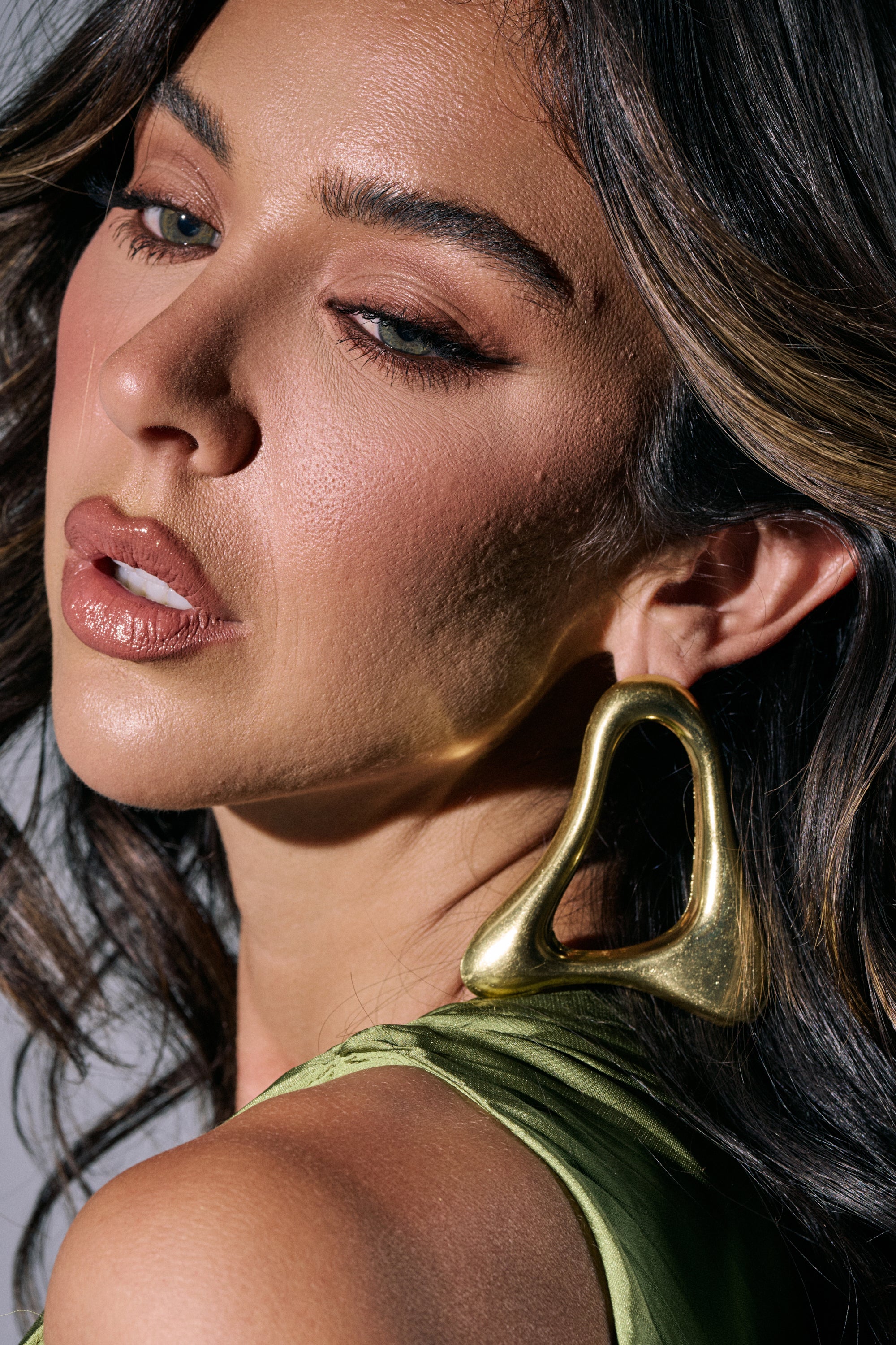 A woman with wavy brown hair and a green top wears the MOD GAL EARRING—sculptural gold statement earrings. Her makeup is natural, featuring defined eyebrows and nude lipstick, as she looks softly to the side. 0