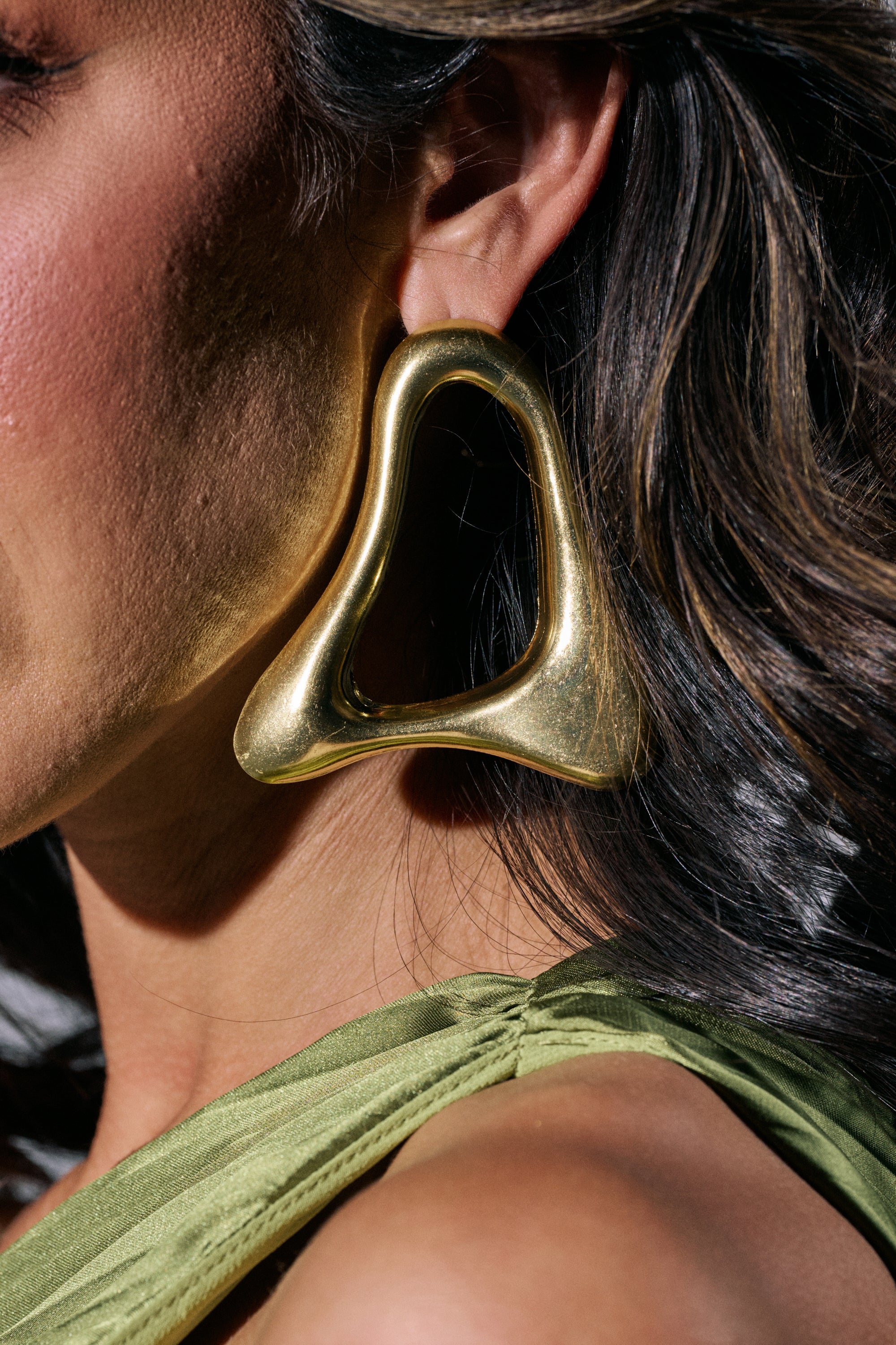 Close-up of a woman's ear wearing the MOD GAL EARRING—large, sculptural gold statement earrings. She has wavy brown hair and wears a gathered-shoulder green top. 1