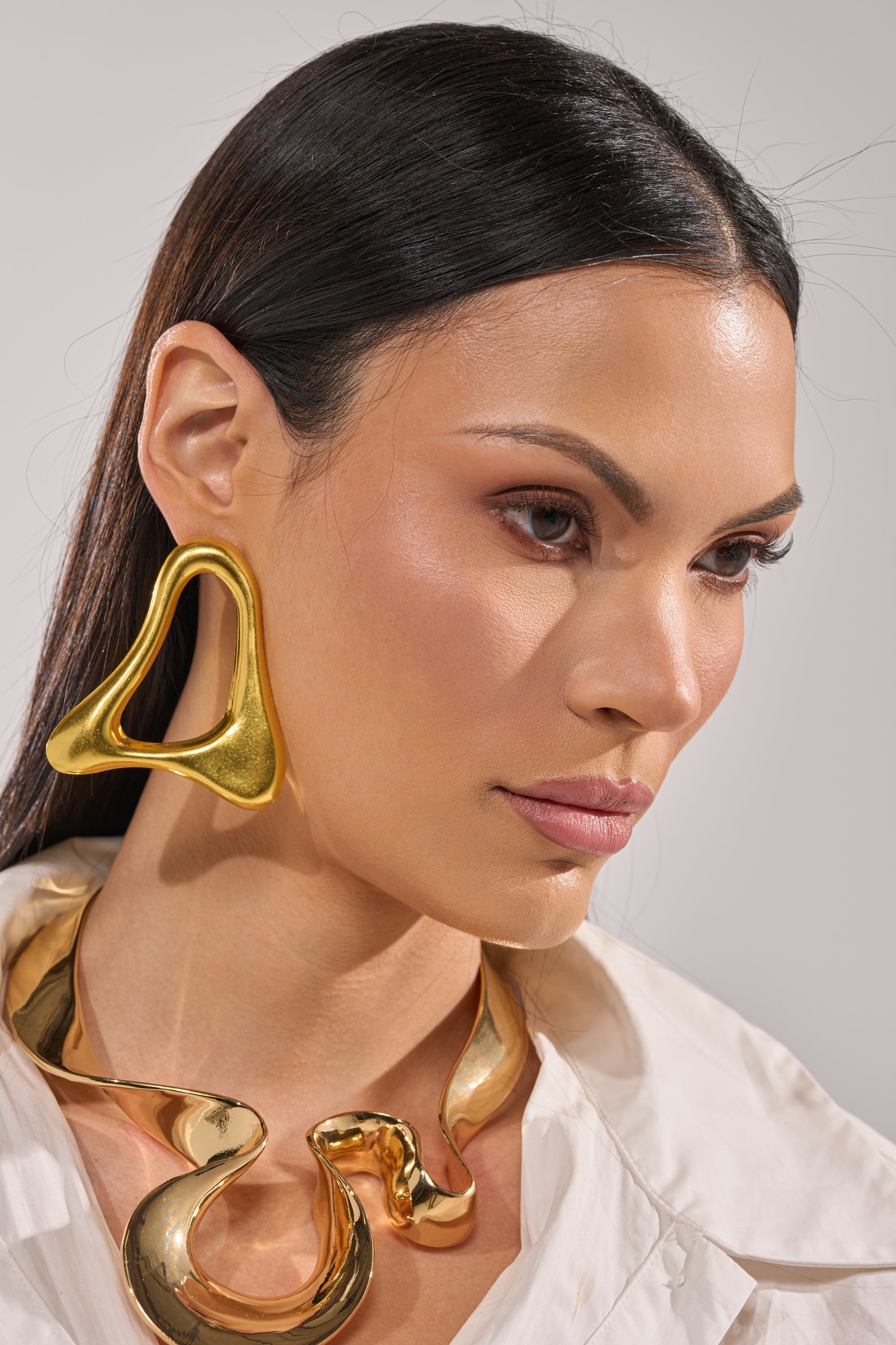 A woman with straight dark hair wears sculptural gold MOD GAL EARRINGS and a chunky gold necklace. Dressed in a white shirt with neutral makeup, she gazes slightly downward and to the side. 5