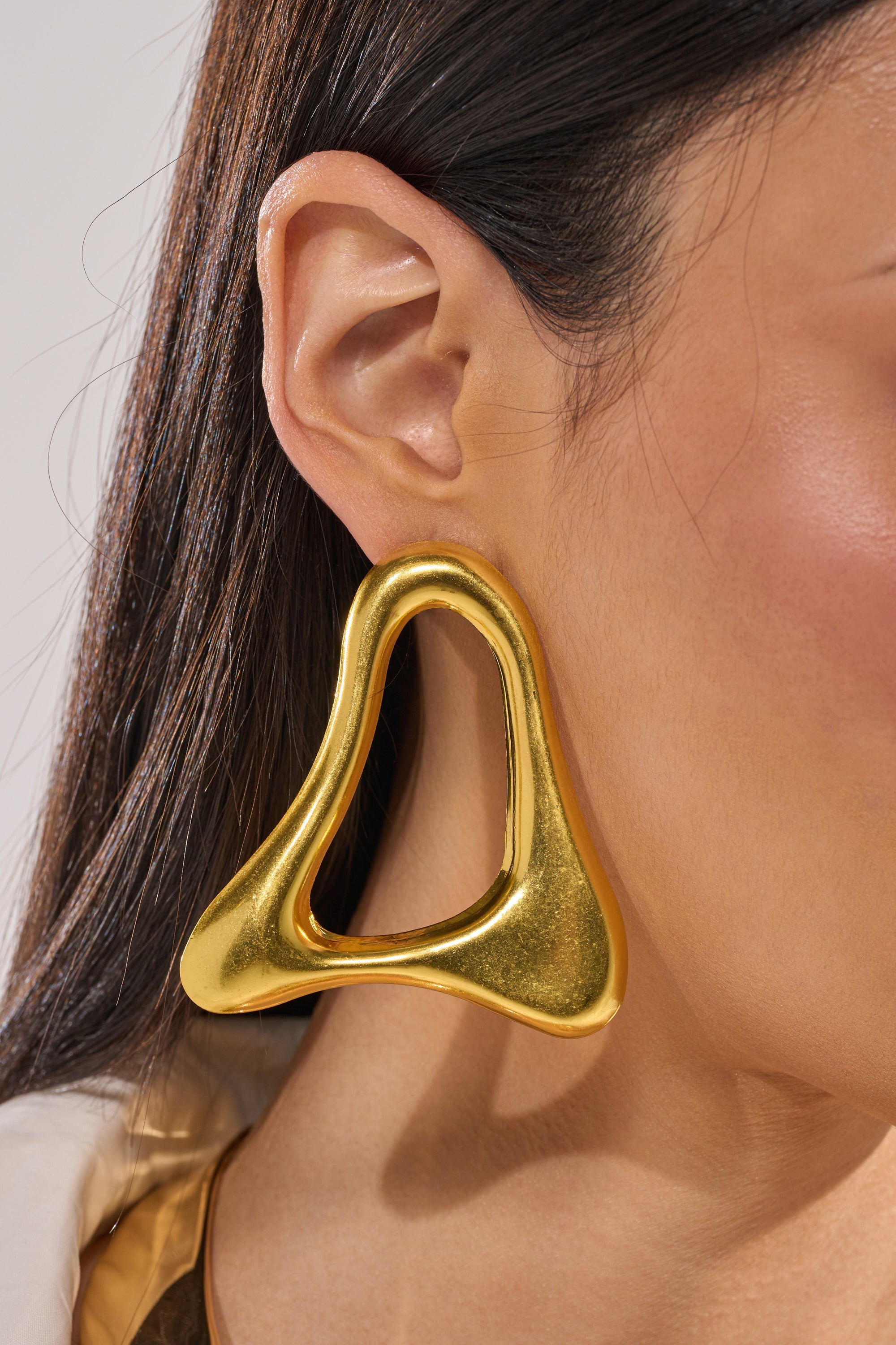 Close-up of a person’s ear wearing the MOD GAL EARRING—a large, shiny gold statement piece with an abstract, hollow center and smooth, rounded edges. The person has straight dark hair and warm-toned skin. 6