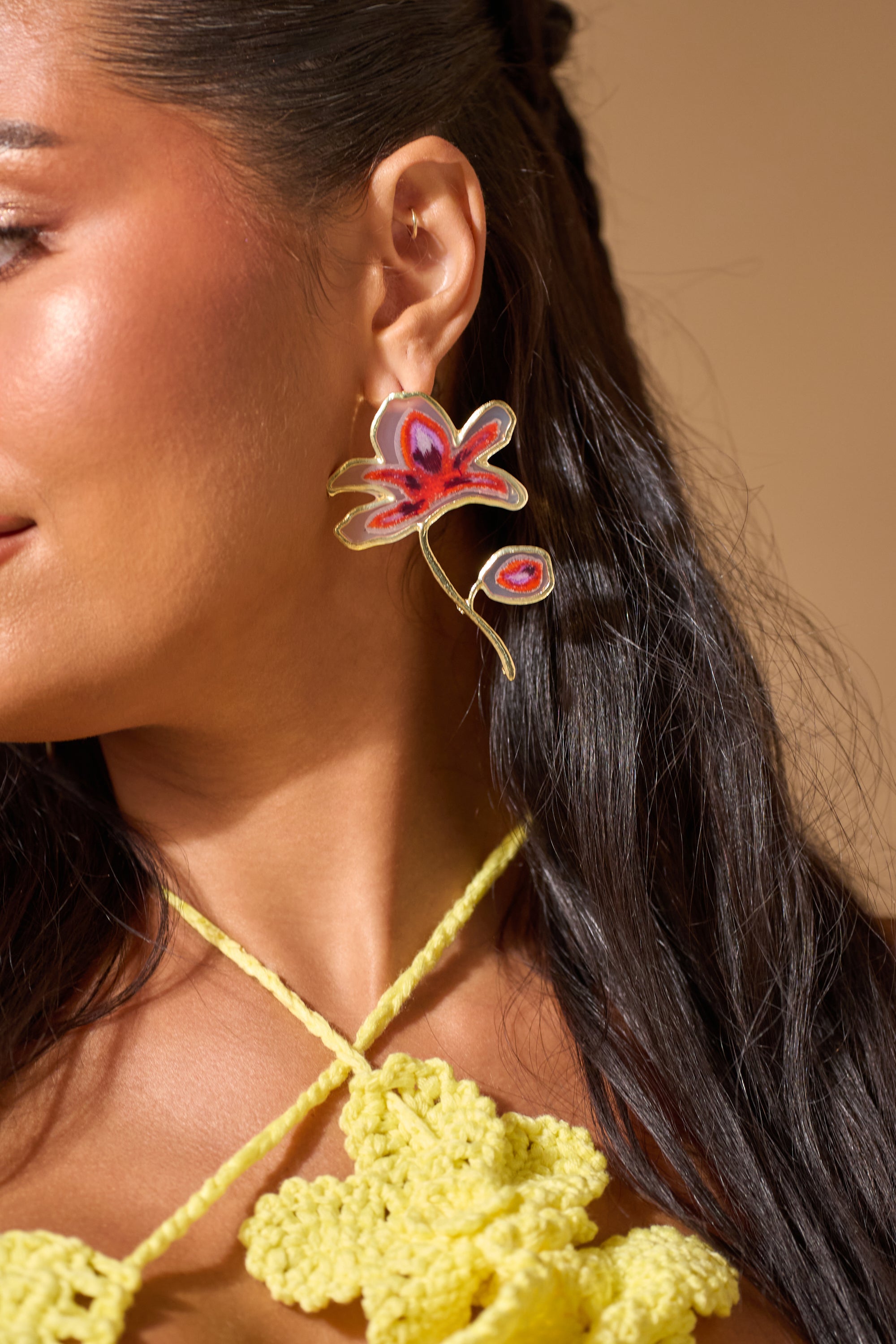 A woman with long dark hair wears the bold IN THE GALLERY EARRING, a red resin floral style, along with a yellow crocheted top featuring floral details, set against a neutral background. 0