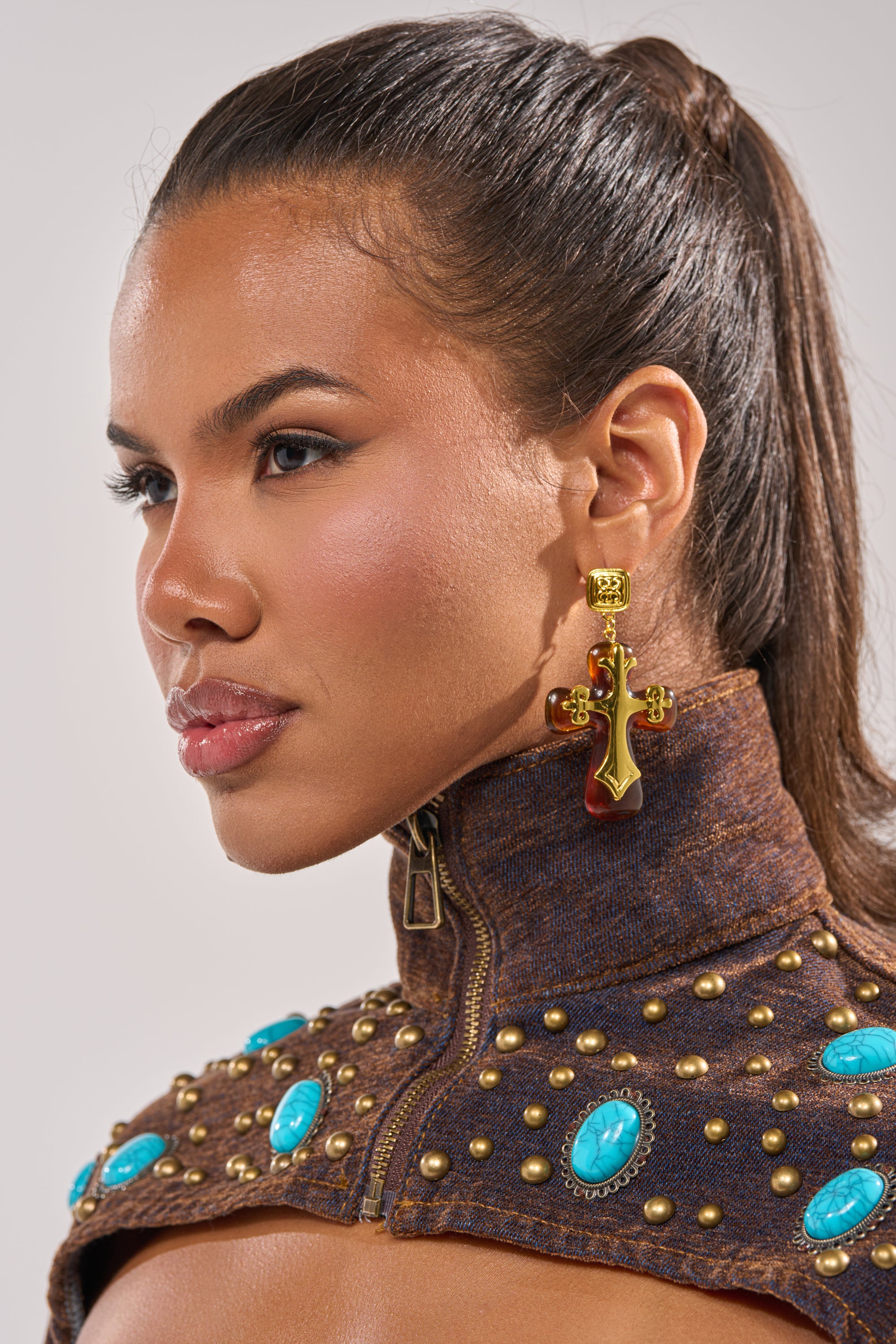 A woman with smooth, glowing skin wears a brown high-collar jacket with turquoise stones and gold studs. Her long hair is pulled back, showcasing the bold CROSS MY HEART EARRING—a large gold cross with striking red accents. 1
