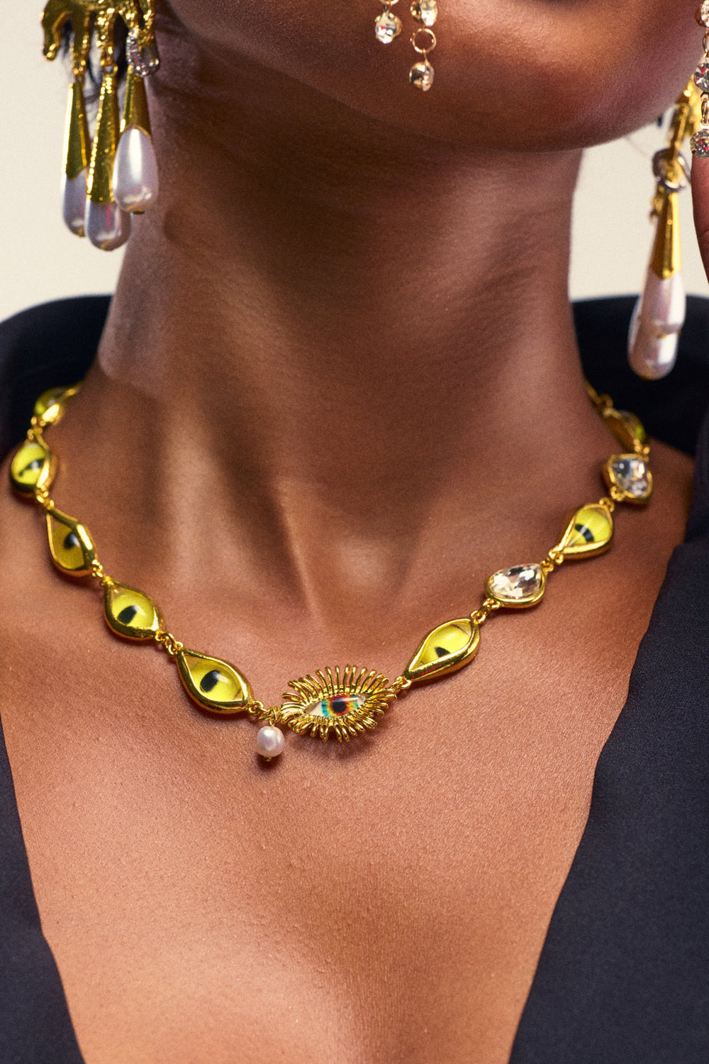 A woman wearing the EYE SEE YOU NECKLACE, a gold statement piece adorned with eye-shaped charms and a central sunburst pendant, complemented by pearl earrings. The necklace features a single dangling pearl—perfect for festival jewelry lovers who appreciate unique details.