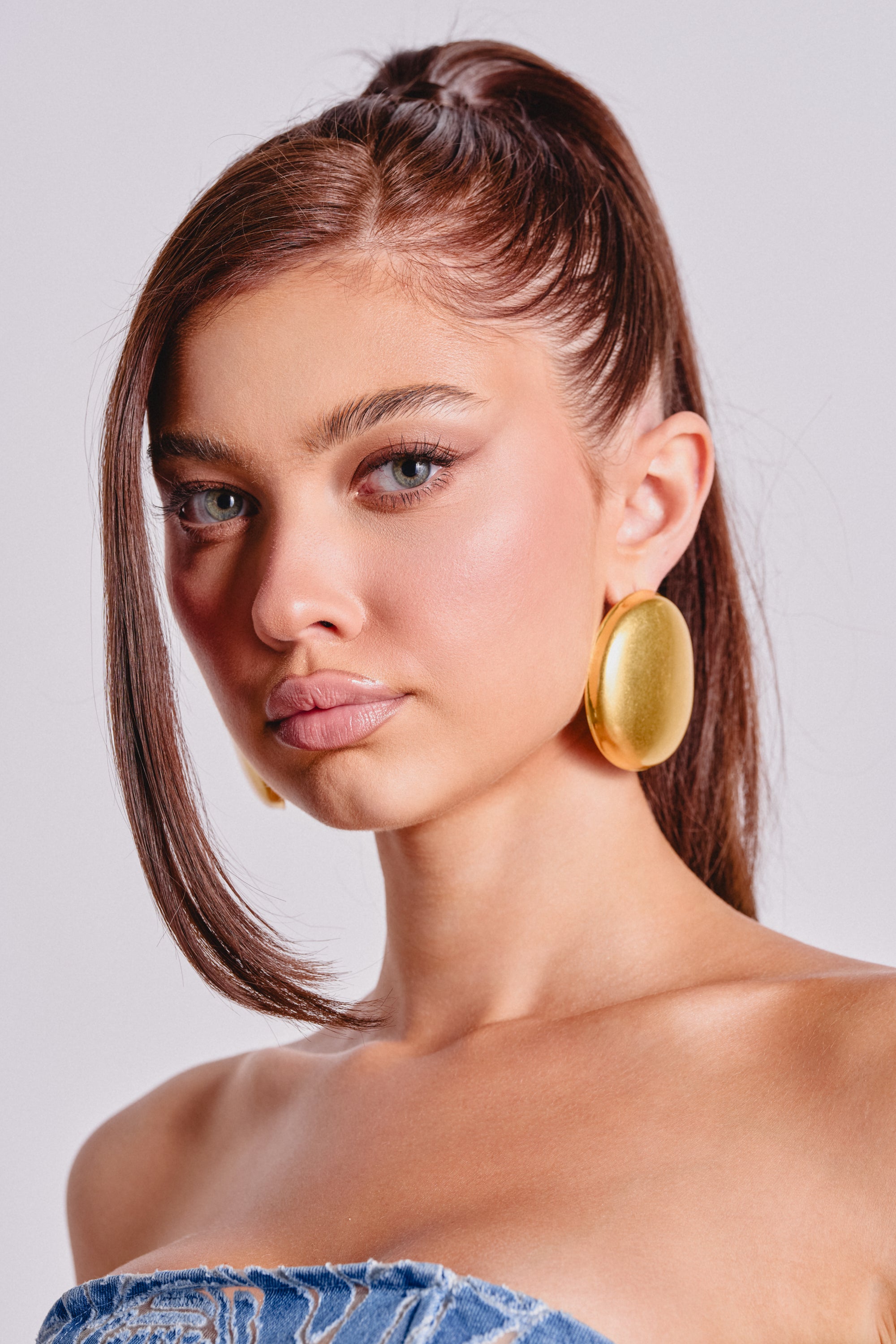 A young woman with straight brown hair in a high ponytail wears the IN TUSCANY EARRING and a denim strapless top, posing against a light background and looking confidently at the camera. 0
