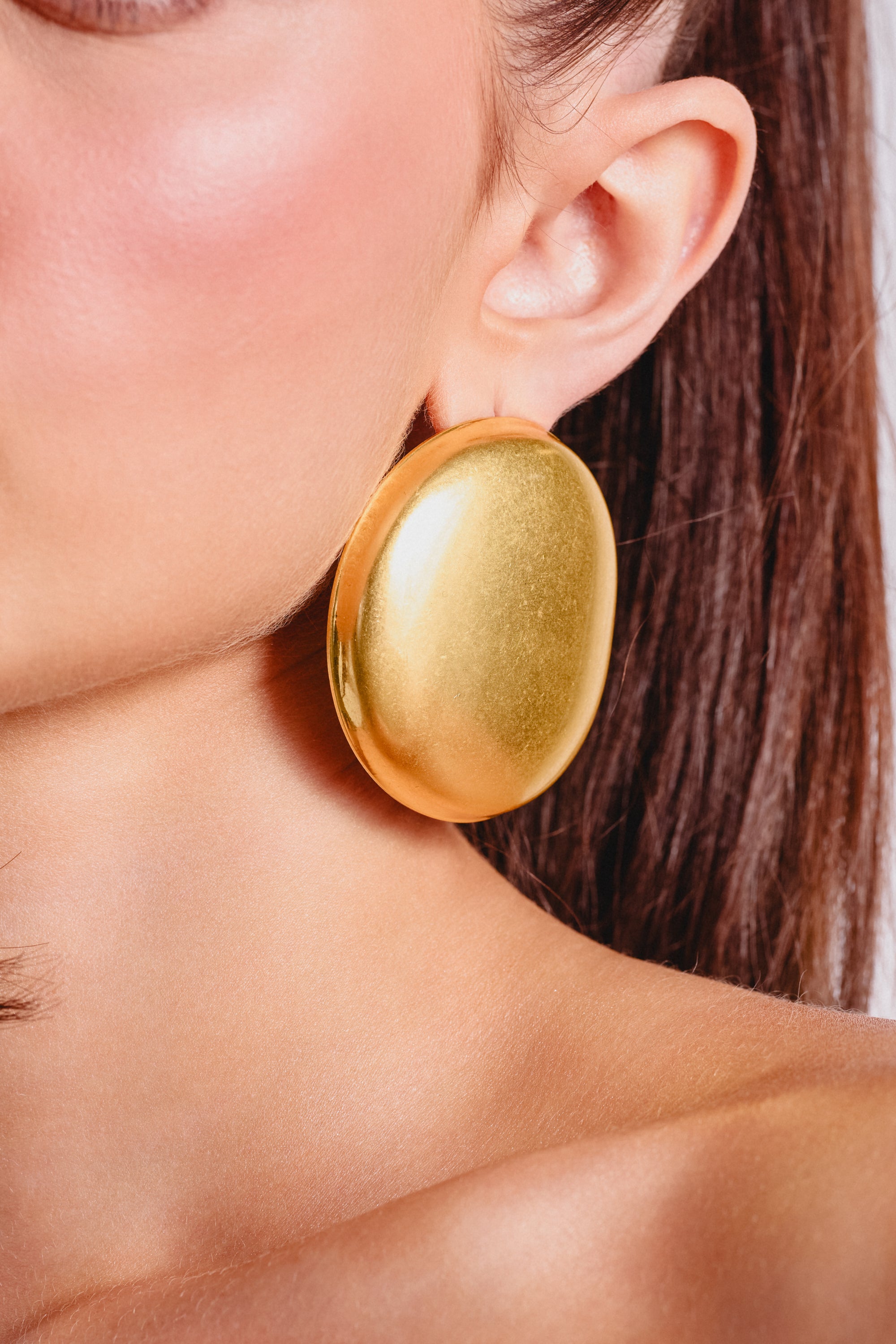 Close-up of a woman's ear wearing the IN TUSCANY EARRING—oversized, sculptural, and shiny gold. Her dark brown hair is pulled back, accentuating her smooth skin with a touch of blush. 1